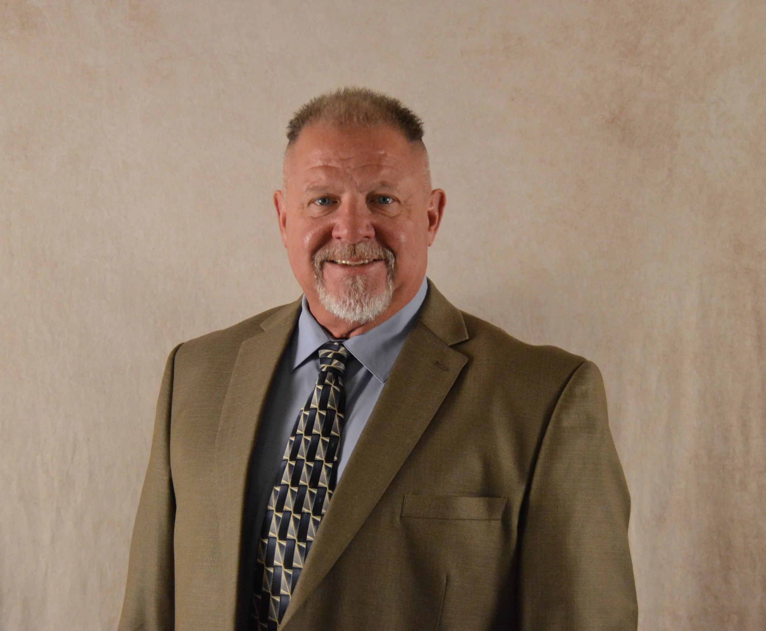A middle-aged man with short, light brown hair and a beard, wearing a brown suit with a light blue shirt and a patterned tie, smiling in front of a beige wall.