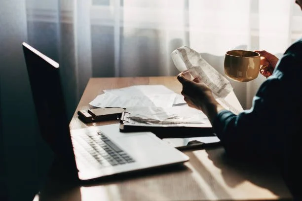 Person holding a paper receipt and a coffee mug at a cluttered desk with a laptop, papers, and a smartphone.