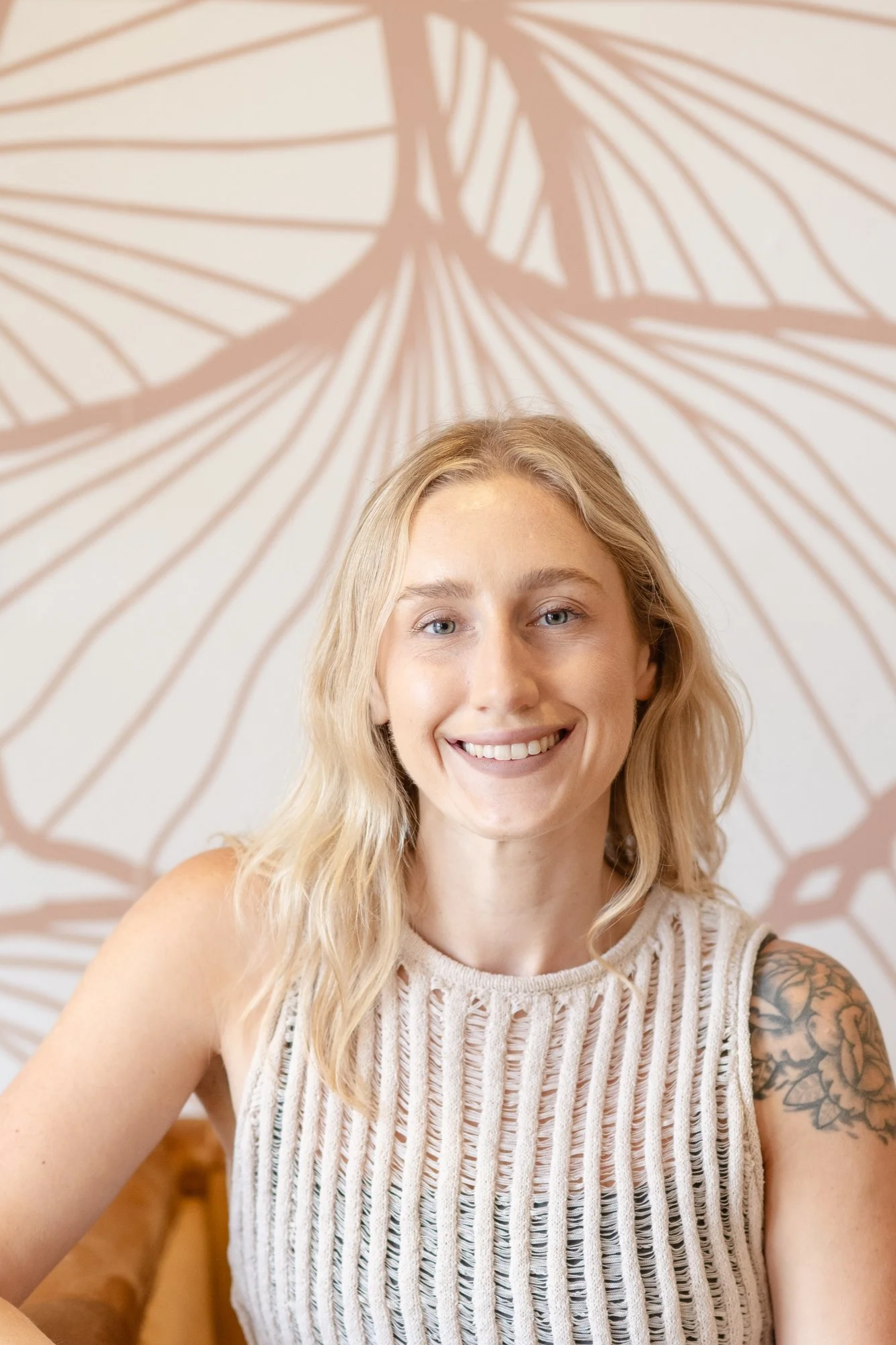 A young woman with blonde hair, smiling and wearing a sleeveless cream-colored knit top, sitting in front of a decorative wall hanging or mural with a geometric pattern behind her.