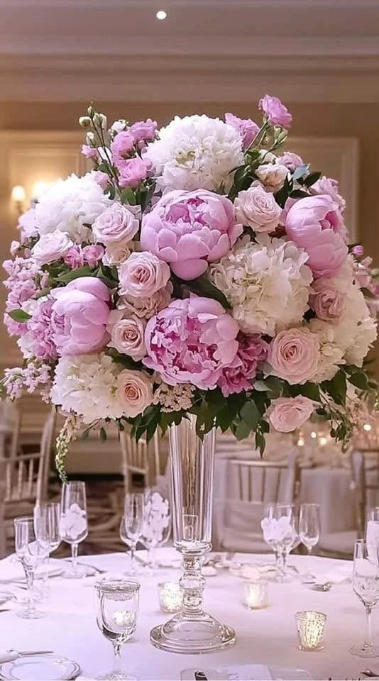 A tall glass vase filled with a large arrangement of pink and white peonies, roses, and hydrangeas on a formal banquet table with glassware and small candle holders in an elegant setting.