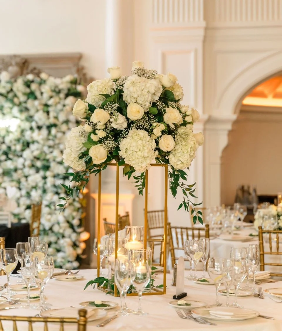 Elegant wedding or event table centerpiece with white roses and hydrangeas on a tall gold stand surrounded by glassware and candles in a decorated room.