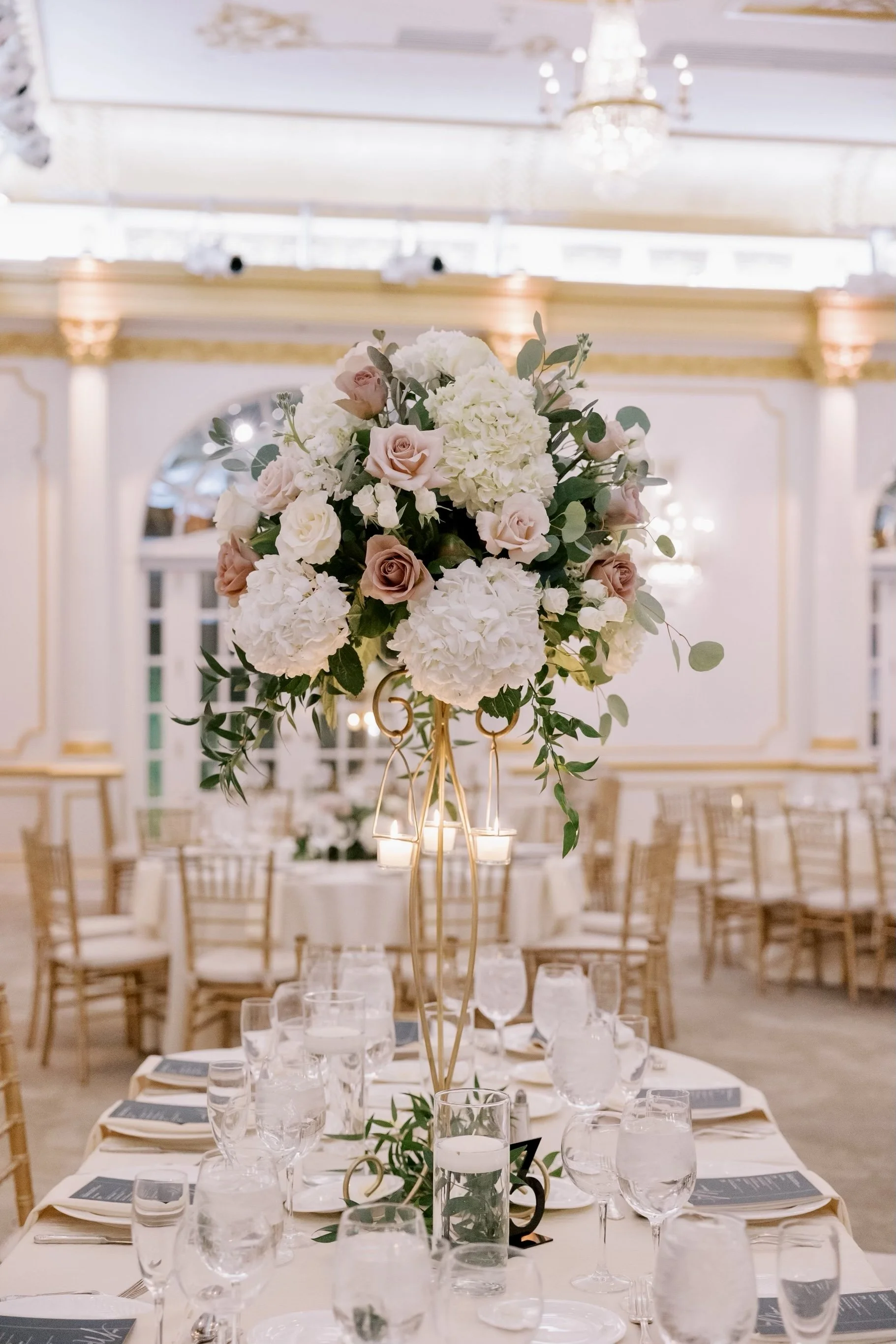 Elegant banquet table with a large floral centerpiece featuring white hydrangeas, blush and cream roses, and eucalyptus, set in an opulent decorated hall with gold accents, chandeliers, and formal place settings with wine glasses and napkins.
