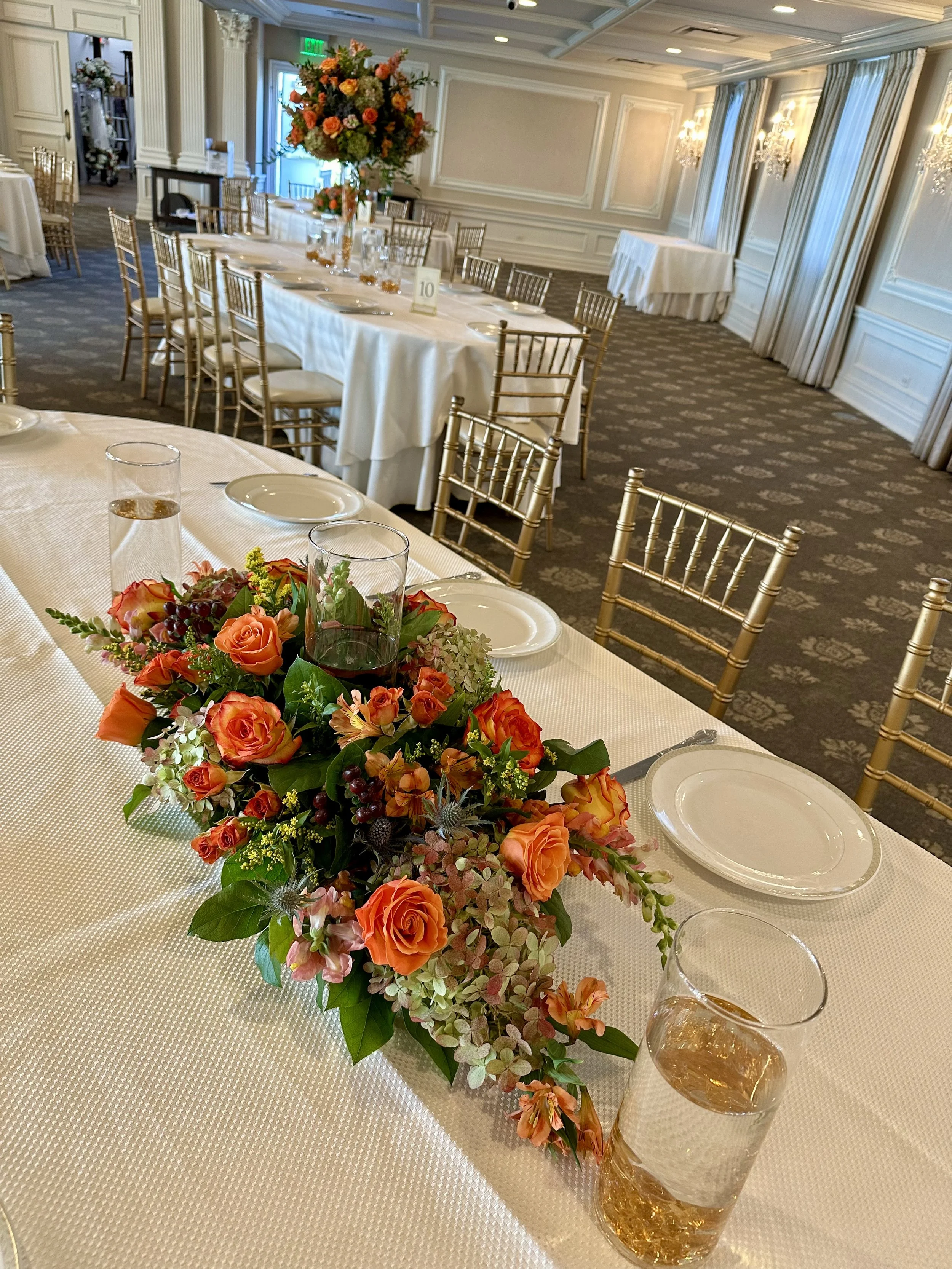 A banquet table decorated with a floral centerpiece featuring orange and pink roses, green foliage, and small filler flowers. The table is set with white plates, glasses, and silverware, and has a white tablecloth. In the background, other tables are