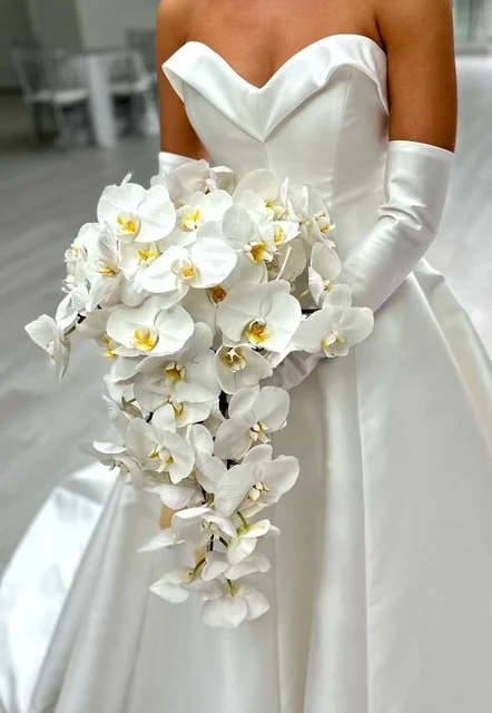A bride wearing a white wedding dress and matching gloves, holding a cascading bouquet of white orchids.