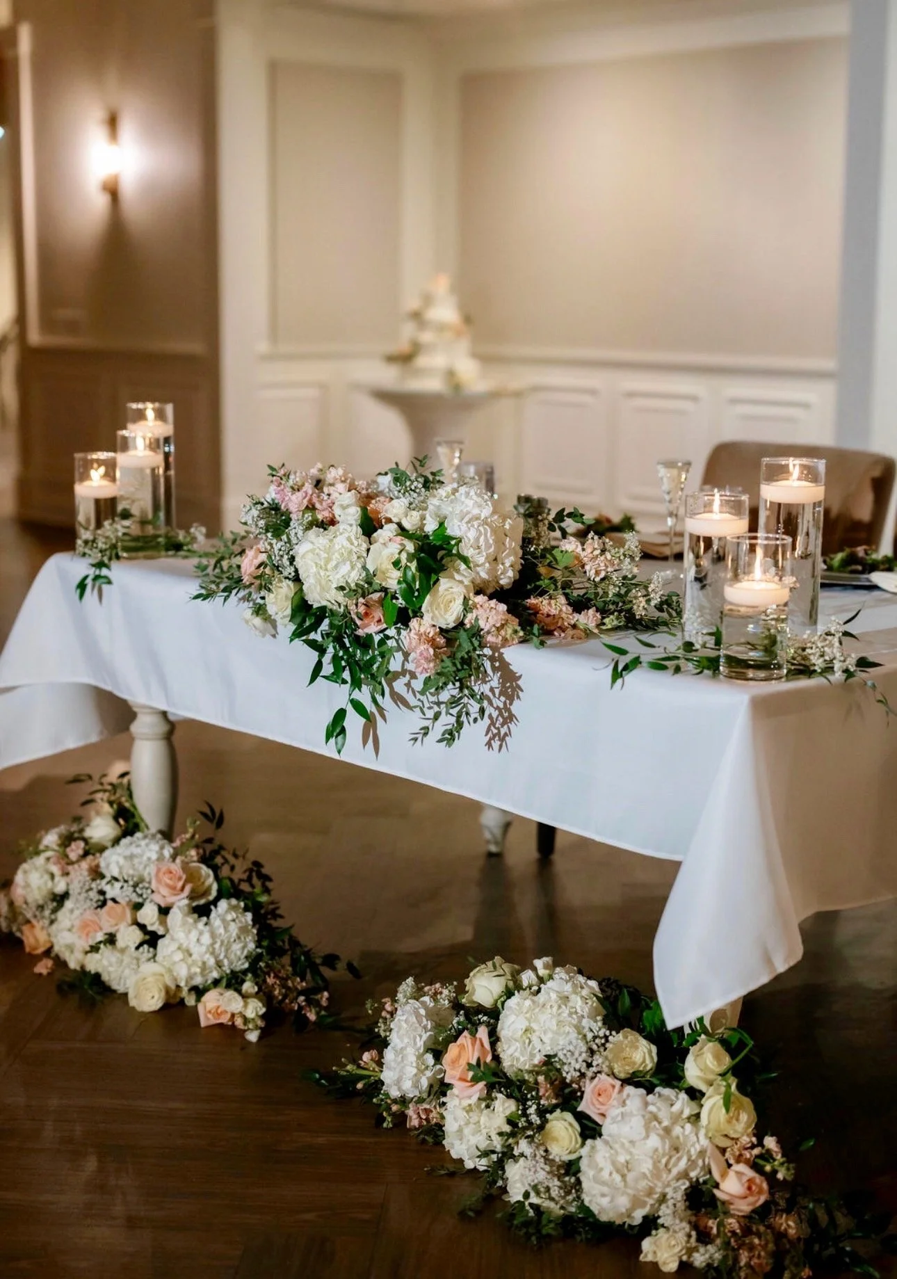 Wedding reception table decorated with a large floral centerpiece of white and light pink flowers, surrounded by candles in tall glass holders, with additional floral arrangements on the floor and a softly lit, elegant room in the background.