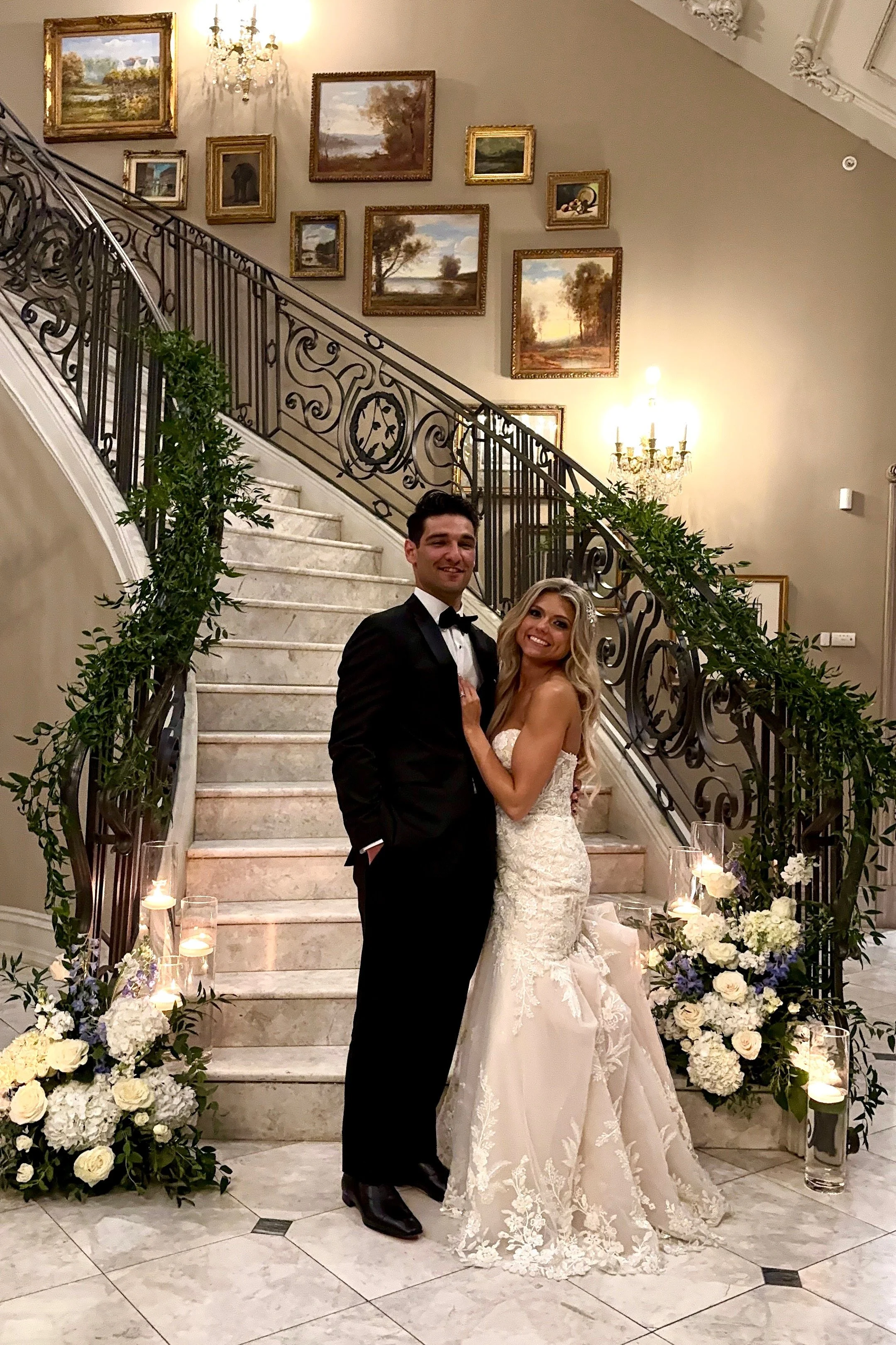 A newlywed couple dressed in wedding attire, standing on a staircase decorated with white and purple flowers, candles, and greenery, inside an elegant, warmly lit room with framed landscape paintings and a chandelier.