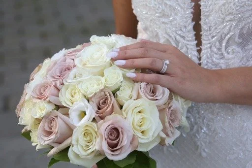A bride holding a bouquet of pink, white, and cream roses, with her hand showing a wedding ring and wearing a wedding dress.