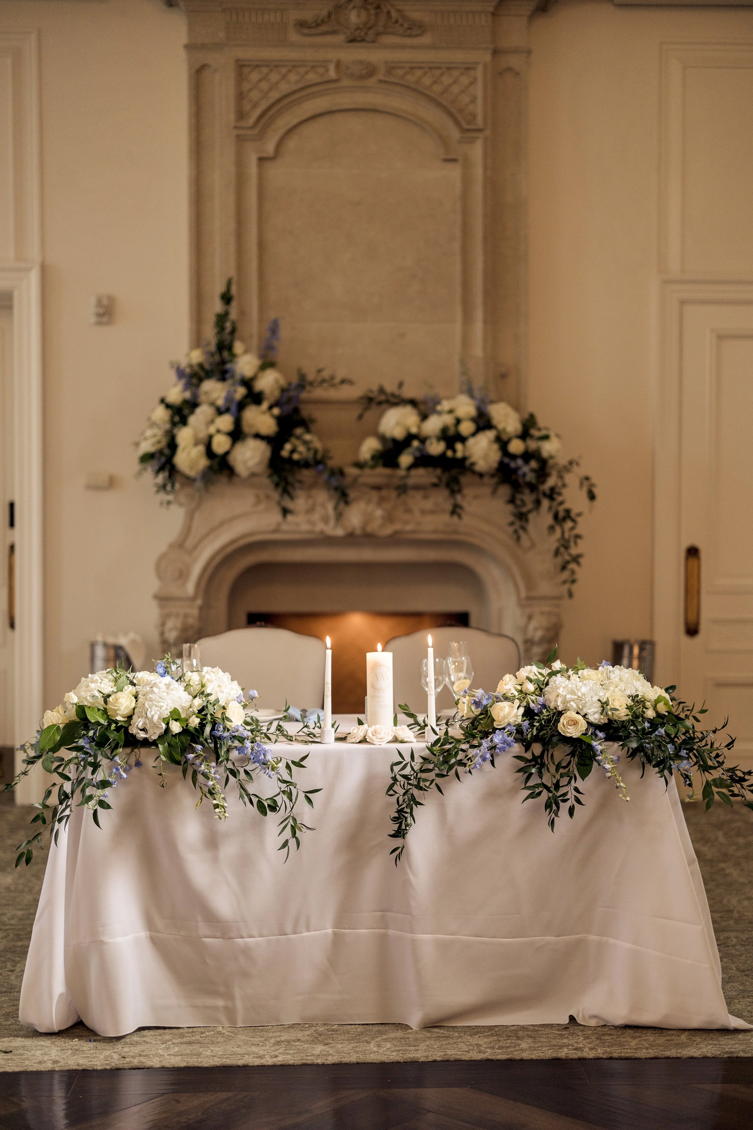 A decorated table for a wedding or special event, featuring a white tablecloth, floral arrangements in white and purple, lit candles, and a cream-colored wall with a fireplace and floral display in the background.
