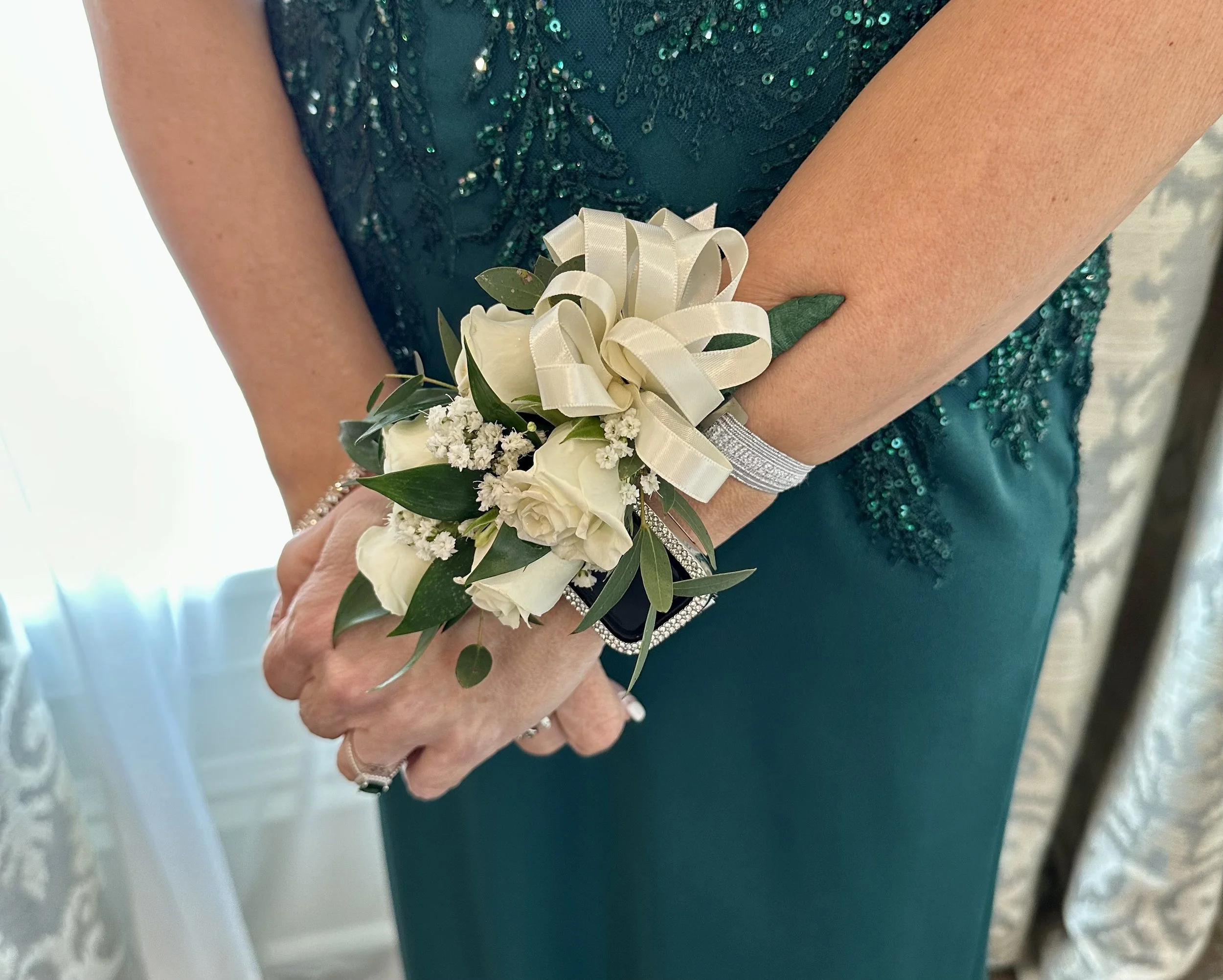 Close-up of a woman’s hand holding a small bouquet of white roses, baby's breath, and green leaves, with a cream-colored ribbon tied around the bouquet, in front of her teal dress with sequins.