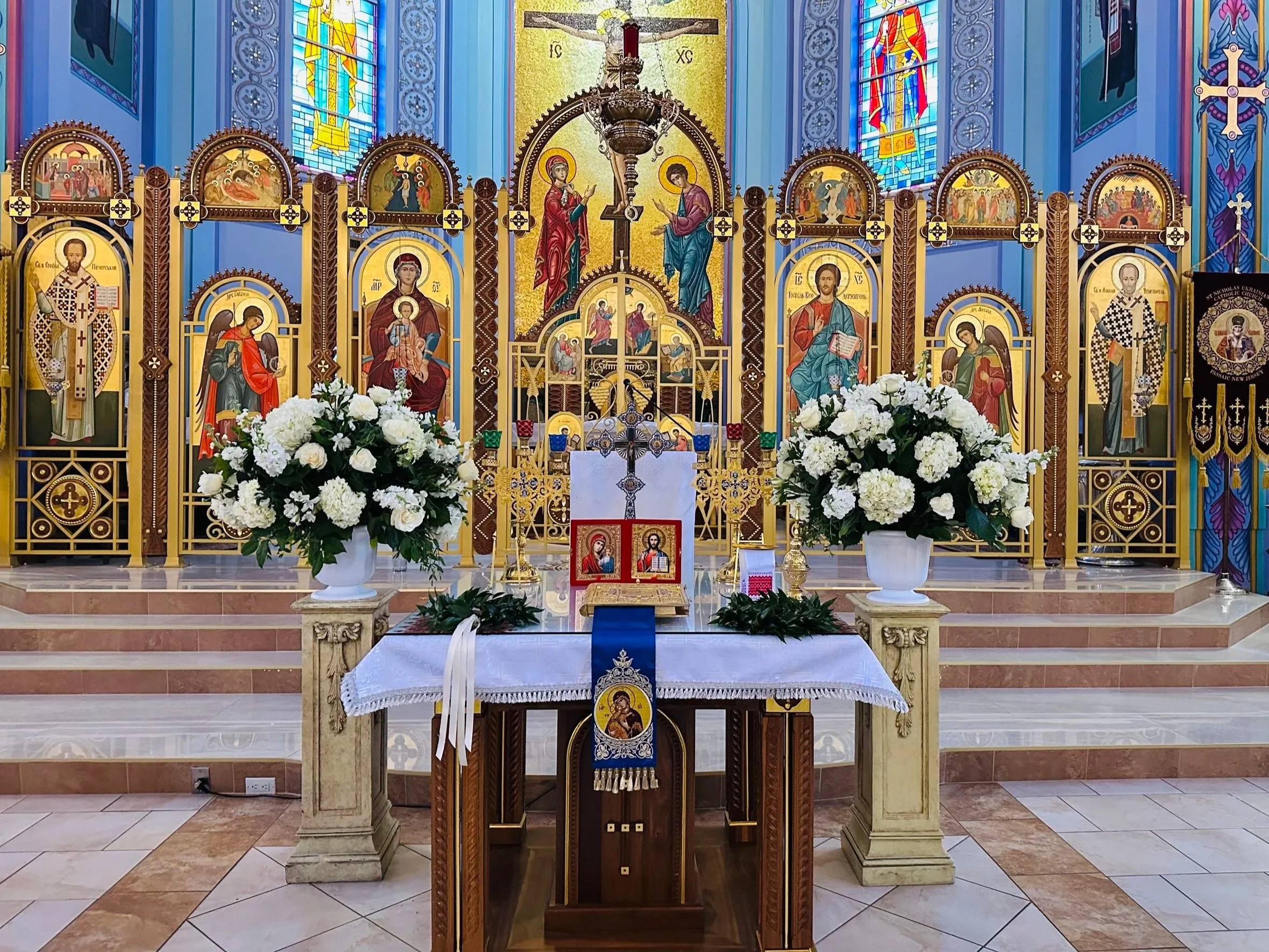 Interior of a church with gold and vibrant colors, featuring religious icons and stained glass windows, with white flowers on the altar.
