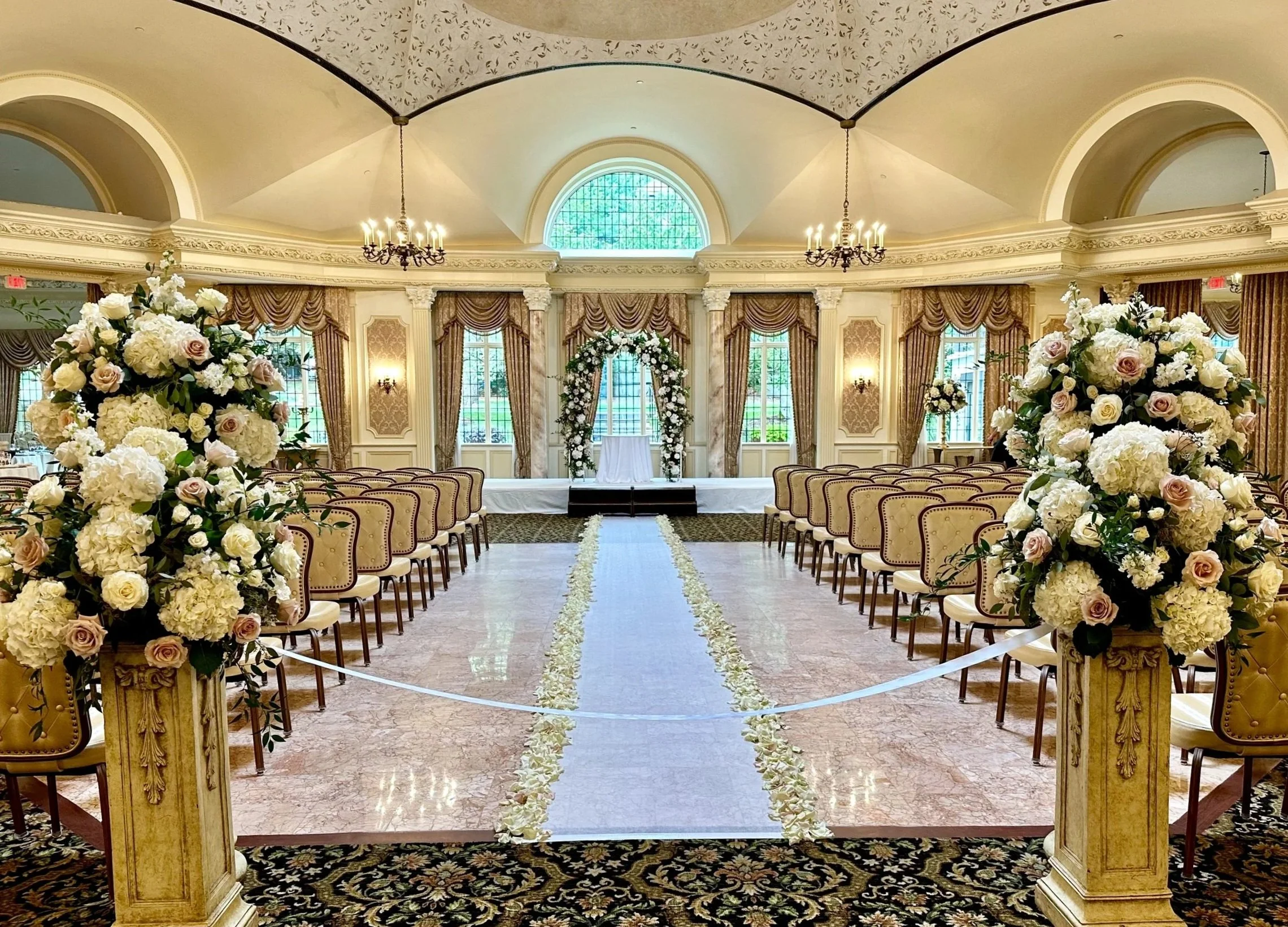 Beautiful wedding ceremony setup in an elegant, classic ballroom decorated with floral arrangements, a floral archway at the front, and rows of chairs for guests.