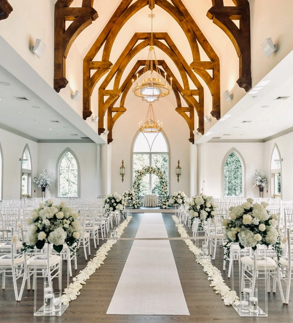 Indoor wedding ceremony setup with white chairs, floral arrangements, a white aisle runner, and a floral arch at the altar inside a church with wooden beams and chandeliers.