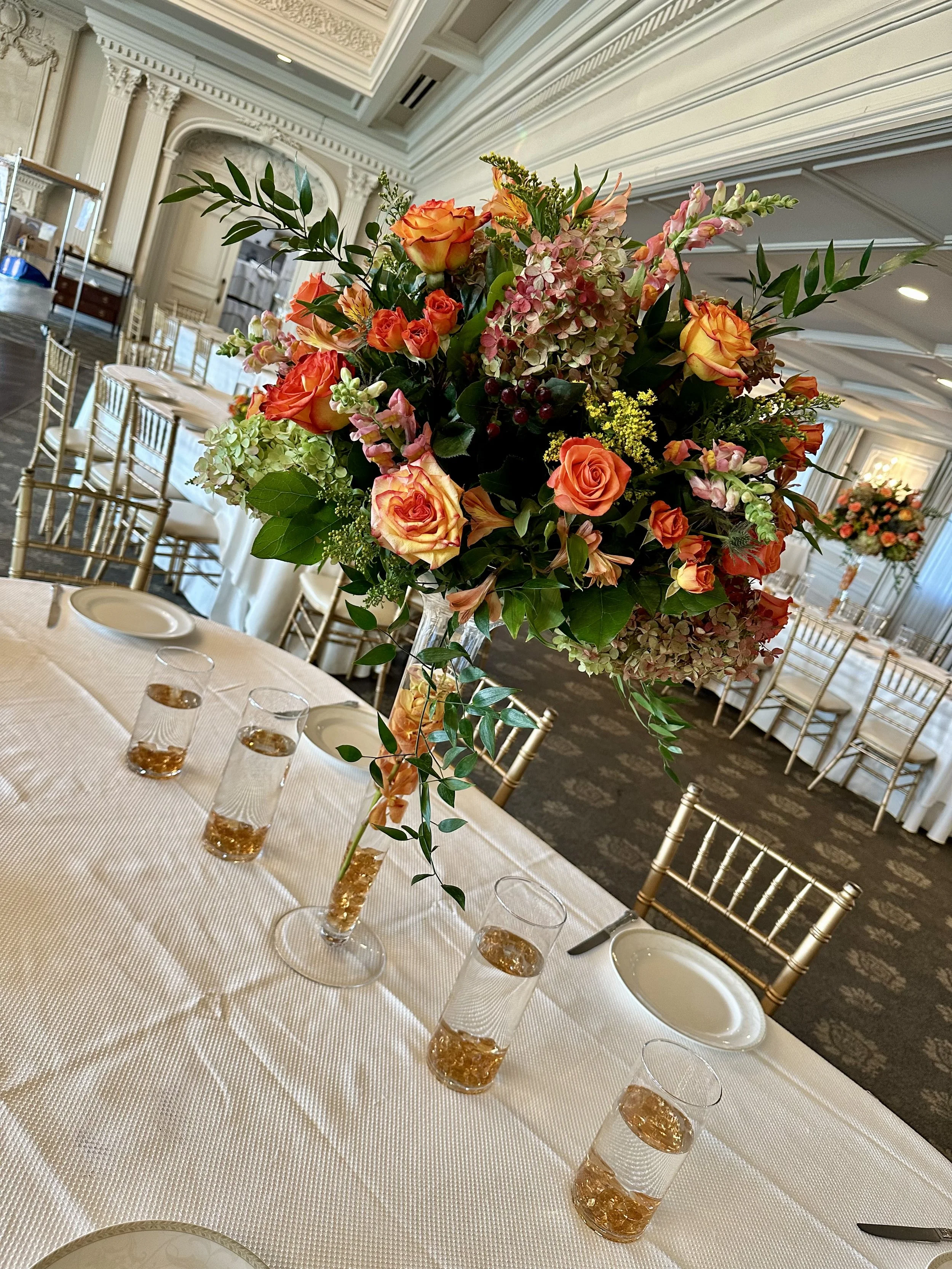 A large floral centerpiece with orange and pink roses, hydrangeas, and greenery on a banquet table at a formal event in an elegant decorated reception hall with gold chairs and white tablecloths.