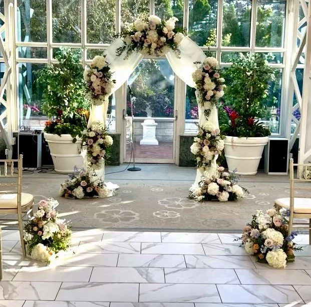 Floral wedding arch with white drapery inside a sunlit room with large windows and potted plants.