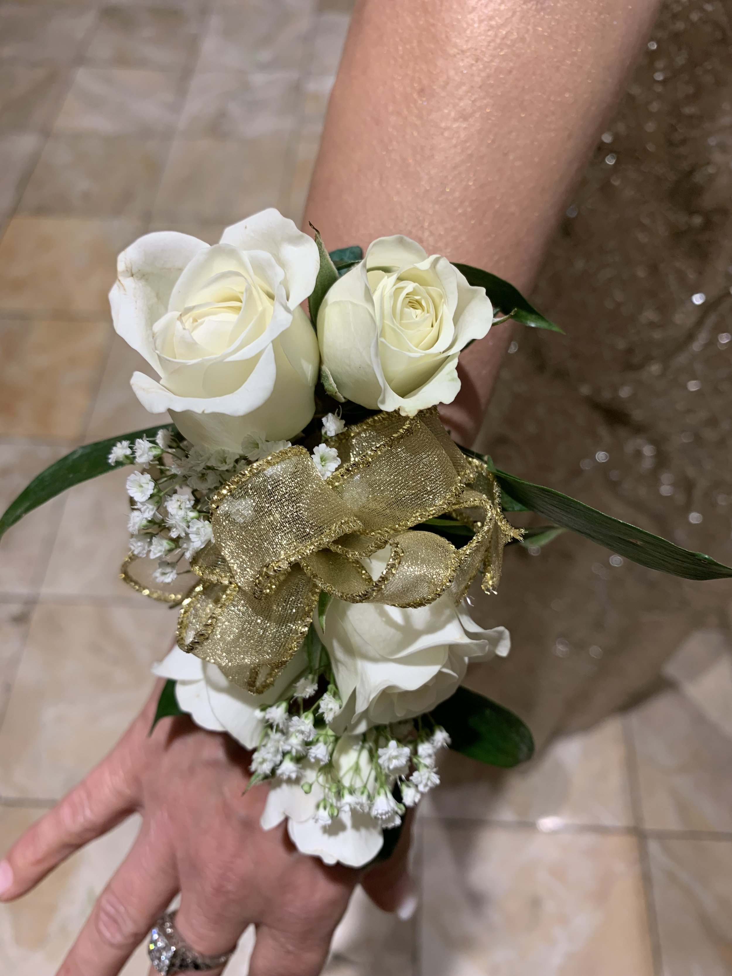 Close-up of a person's hand holding a boutonniere with white roses, small white filler flowers, green leaves, and a gold ribbon, against a blurred tiled floor background.