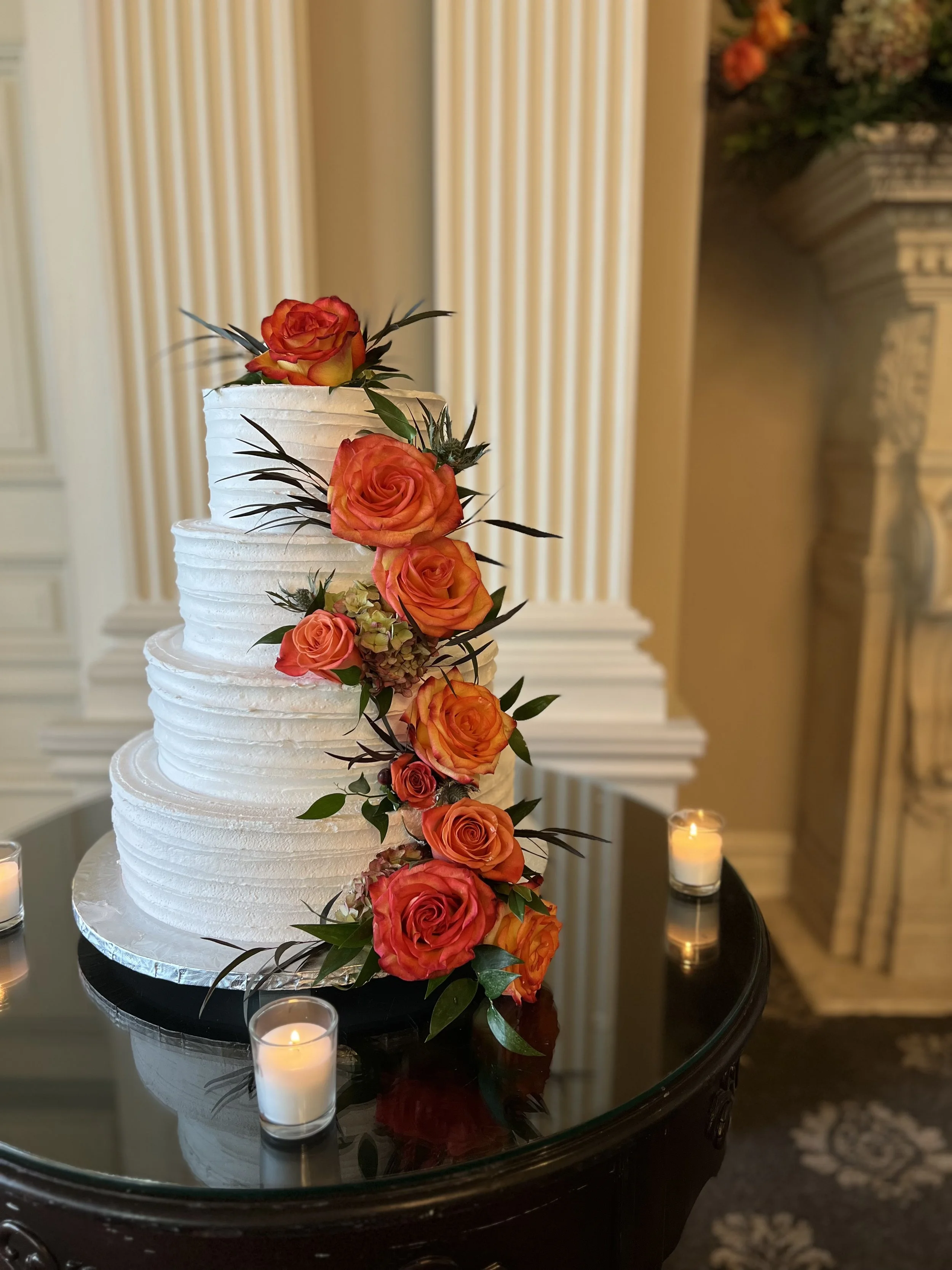 A three-tiered white wedding cake decorated with orange and pink roses and green leaves, on a black glass table with small lit candles, in an elegant interior setting