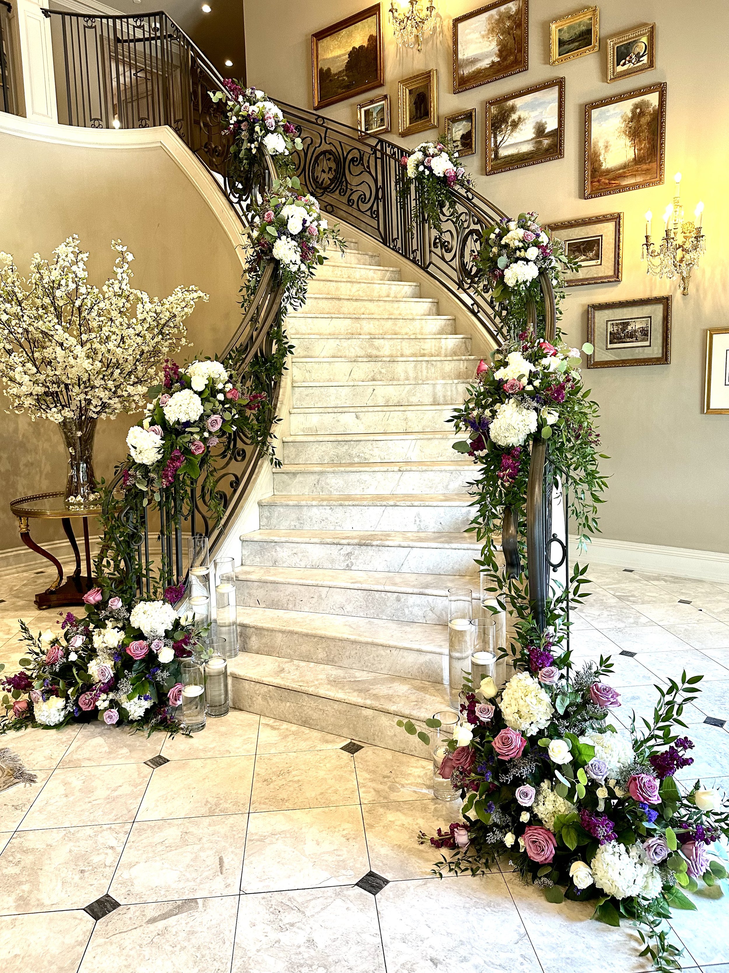 Elegant stairway decorated with floral arrangements of white, purple, and pink roses and hydrangeas, accented with greenery, in a well-lit interior space with artwork on the walls and a chandelier overhead.