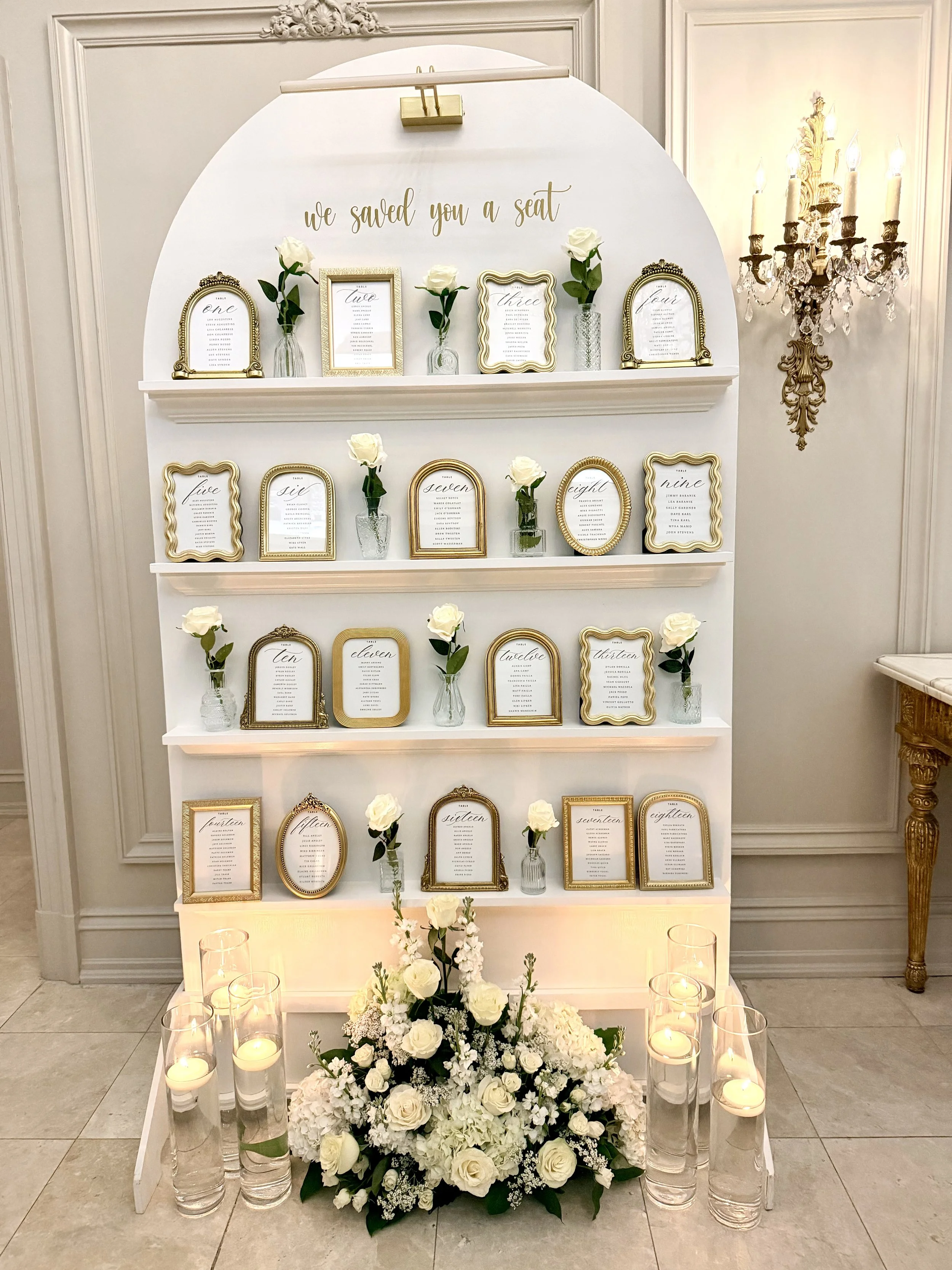 Elegant wedding seating chart display with framed cards on white shelves, white roses in glass vases, a floral arrangement with white roses and hydrangeas, surrounded by candles in glass holders, in a luxurious setting with ornate wall decor and a ch