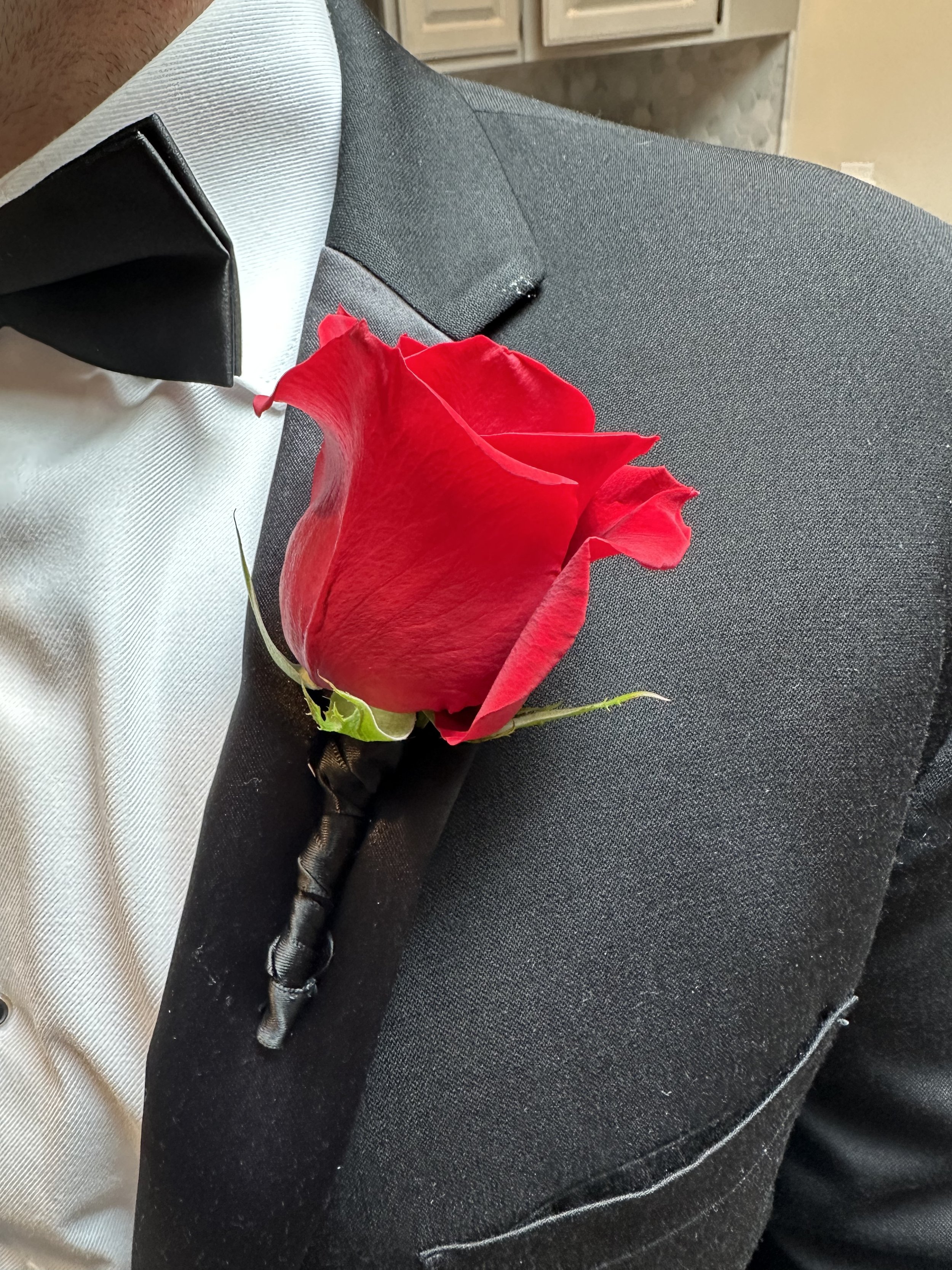A close-up of a person's chest showing a red rose boutonniere pinned to a dark gray suit jacket and white shirt.