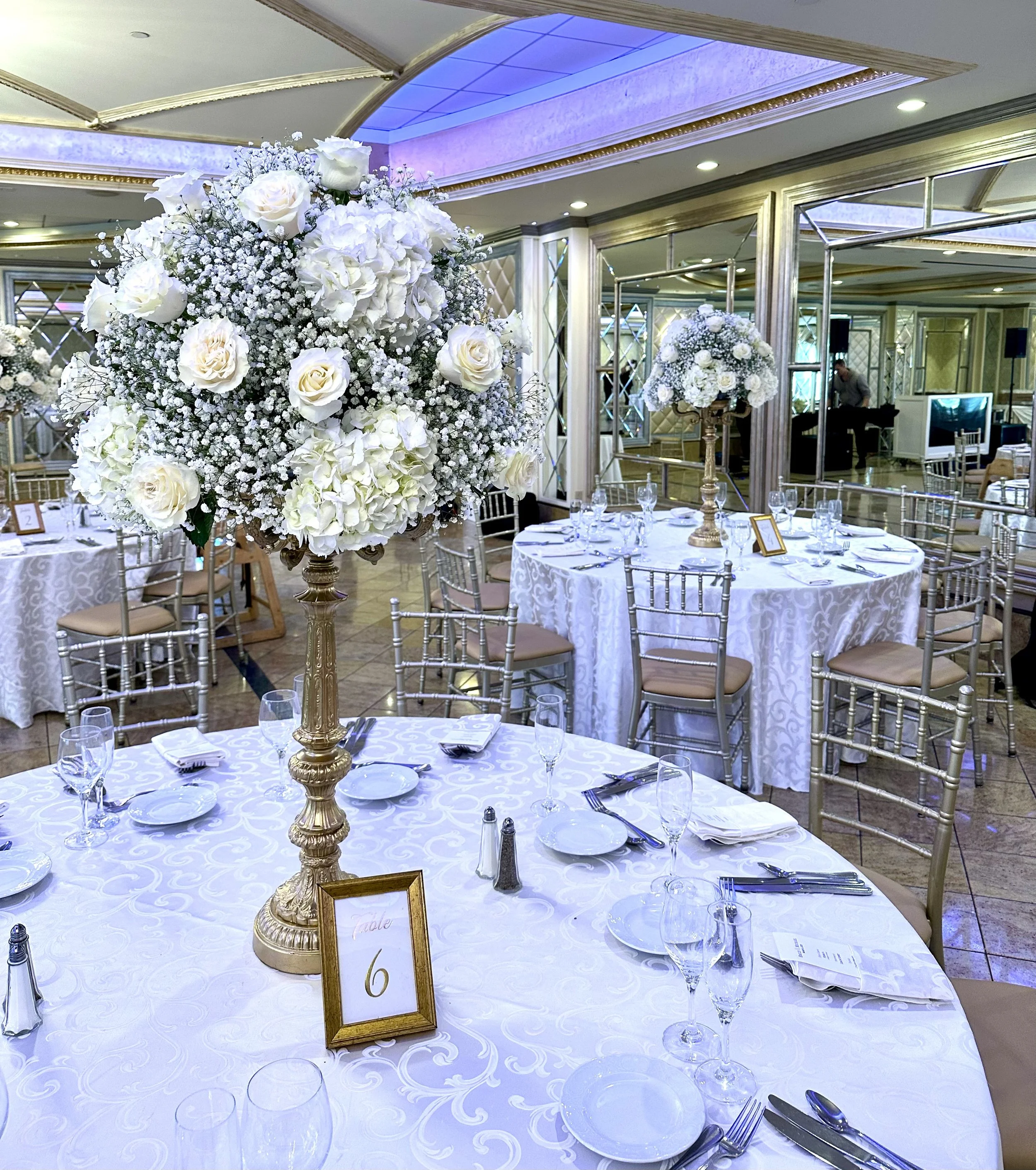 Elegant banquet table set with white tablecloth, glassware, silverware, and a large floral centerpiece featuring white roses and baby's breath in a gold stand, in a decorated hall with mirrors and ceiling lighting.