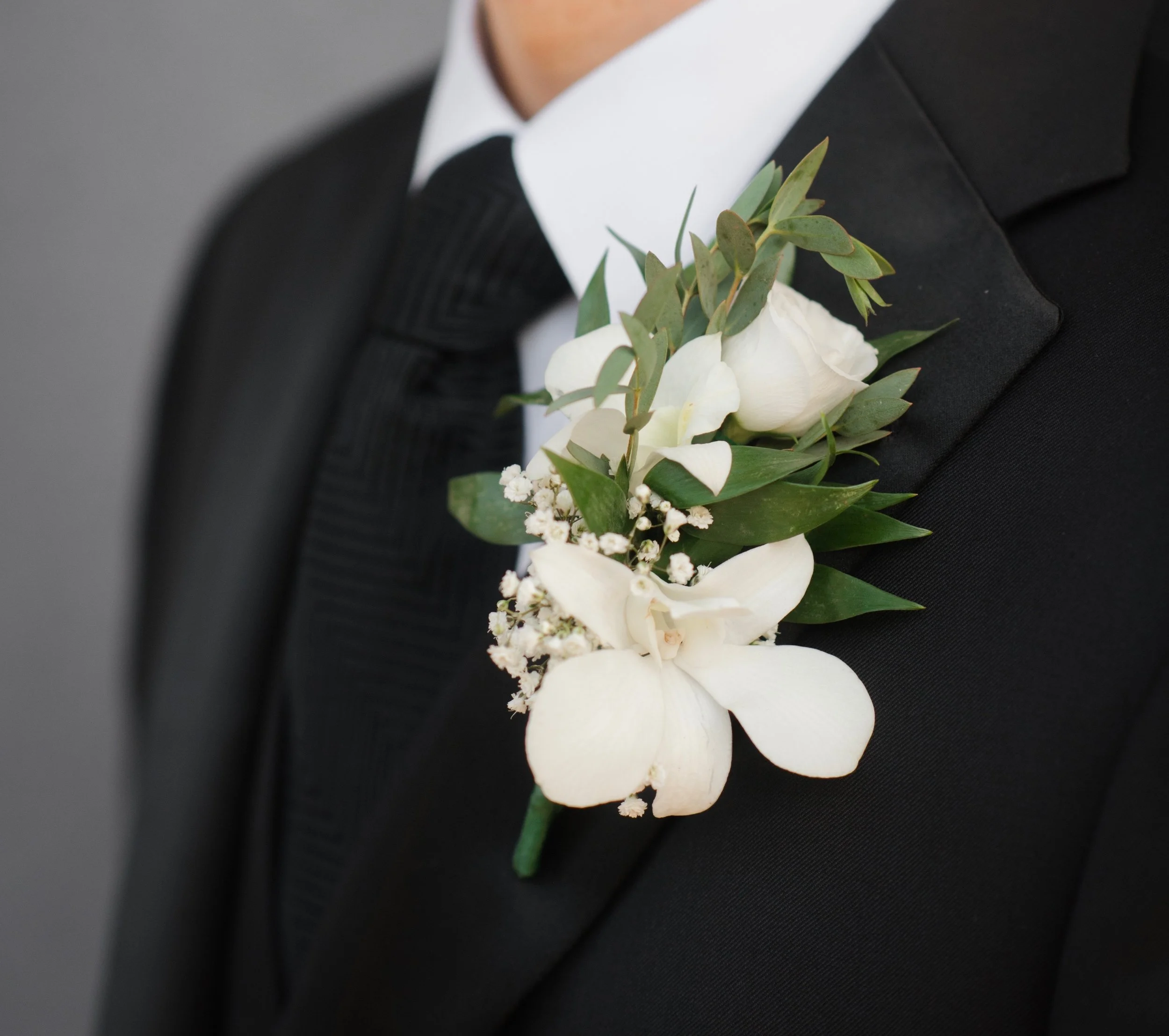 Close-up of a person wearing a black suit and tie with a white shirt, decorated with a boutonnière made of white flowers and green leaves, likely for a formal event or wedding.