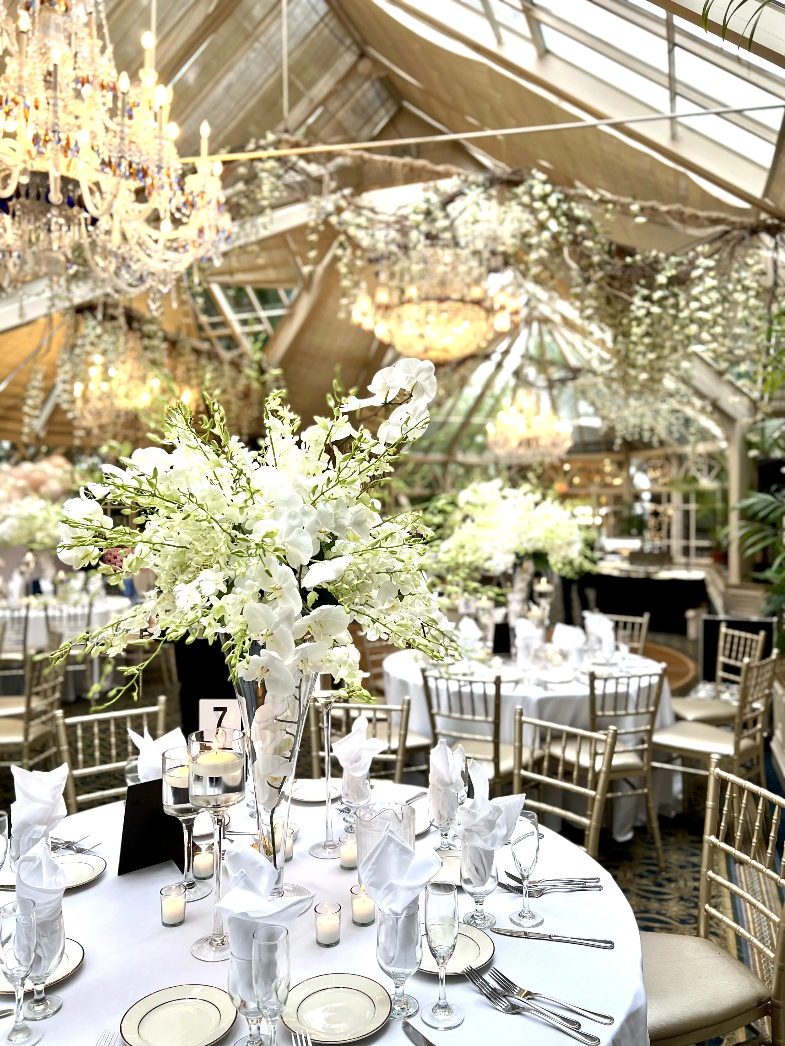 Elegant banquet hall with white tablecloths, floral centerpieces, and lit candles. Round tables surrounded by gold chairs. Decorated with chandeliers and floral arrangements hanging from the ceiling.