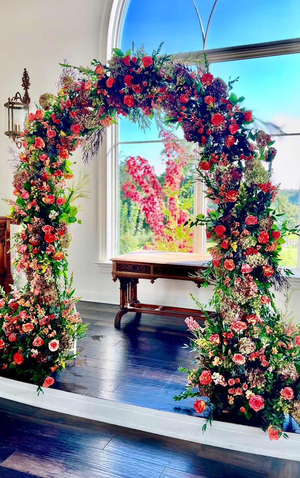 Colorful floral arch with pink roses and various flowers set inside a room with large window and dark wood flooring.