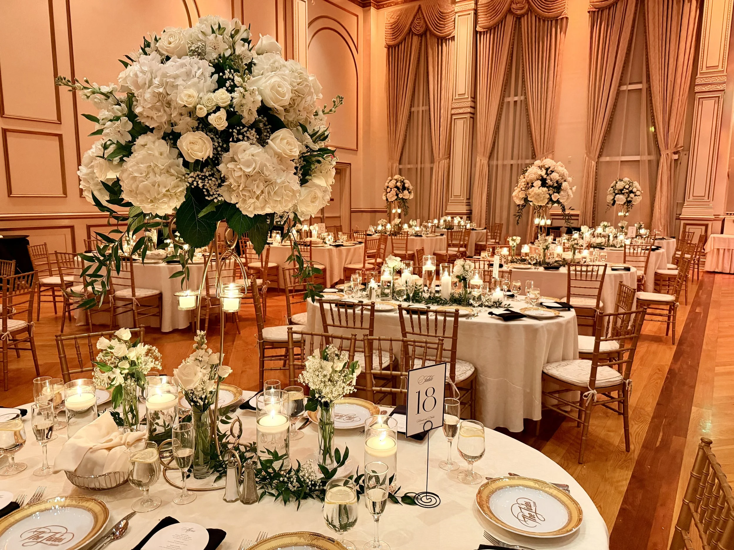 Elegant banquet hall with large floral centerpiece featuring white roses and hydrangeas, surrounded by lit candles and table settings with gold-rimmed plates and glasses, in a room with tall windows and coral-colored curtains.