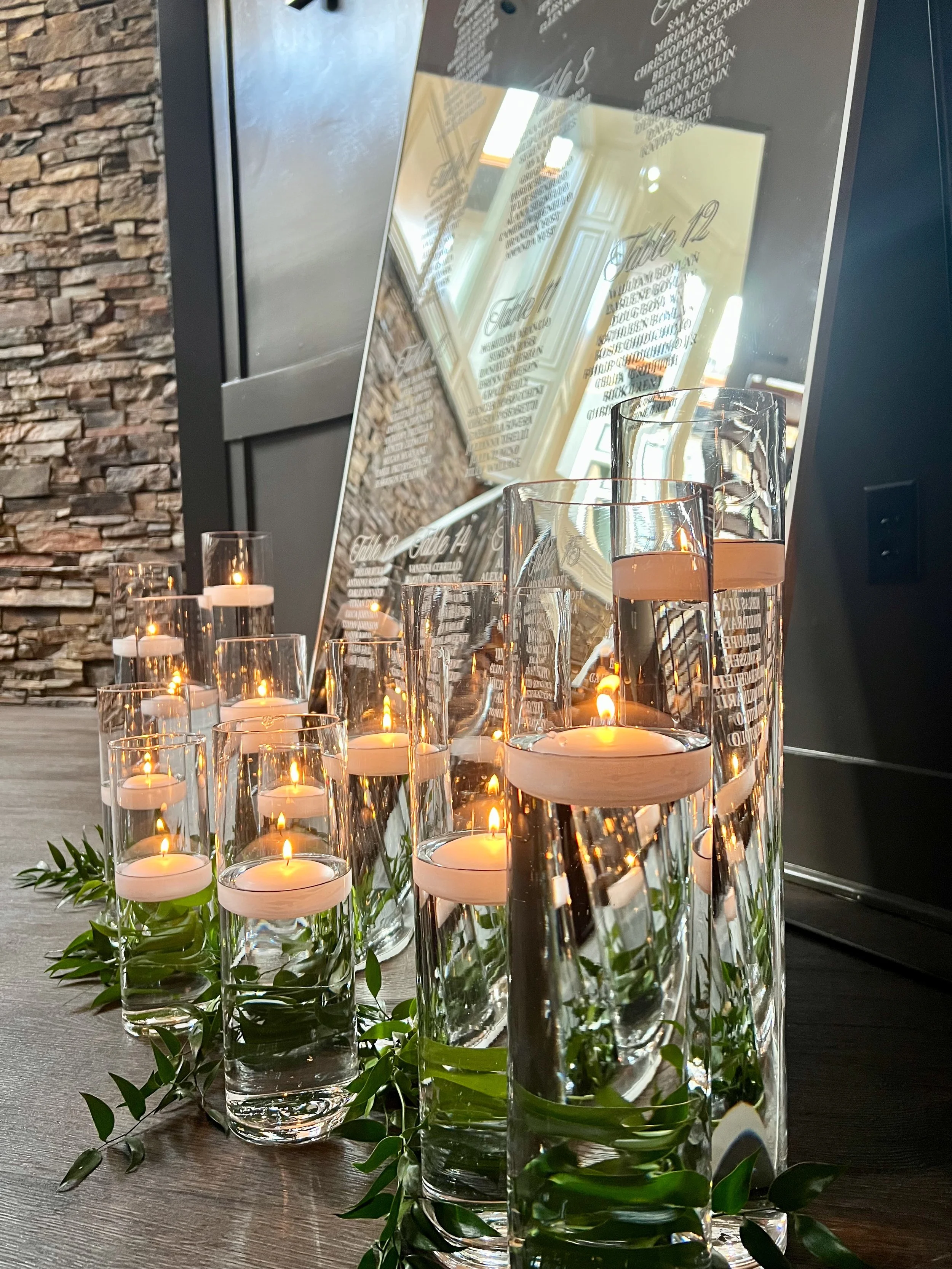 Multiple lit candles in tall glass holders with green leaves at their base, placed on a wooden table in front of a reflective menu board in a restaurant or cafe.