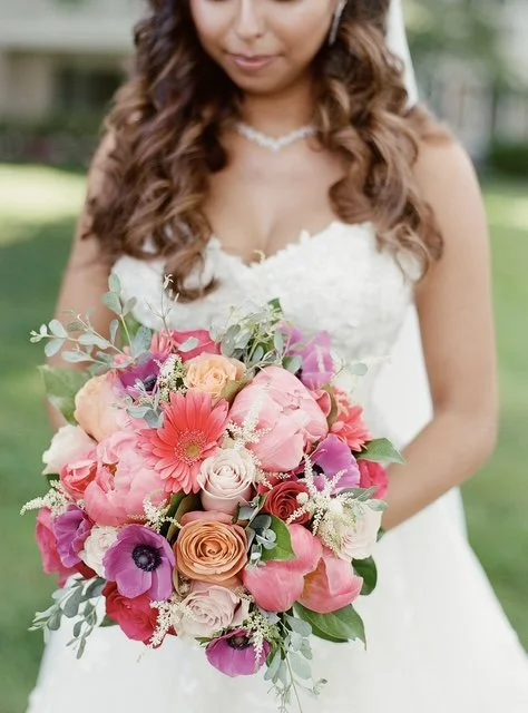 A bride in a white wedding dress holding a bouquet of pink, peach, purple, and white flowers with green foliage, outdoors.