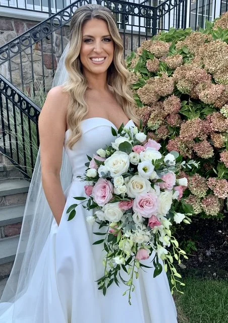 A bride in a white wedding gown holding a large bouquet of white and pink roses, standing outdoors near a metal staircase and pink hydrangea bushes.