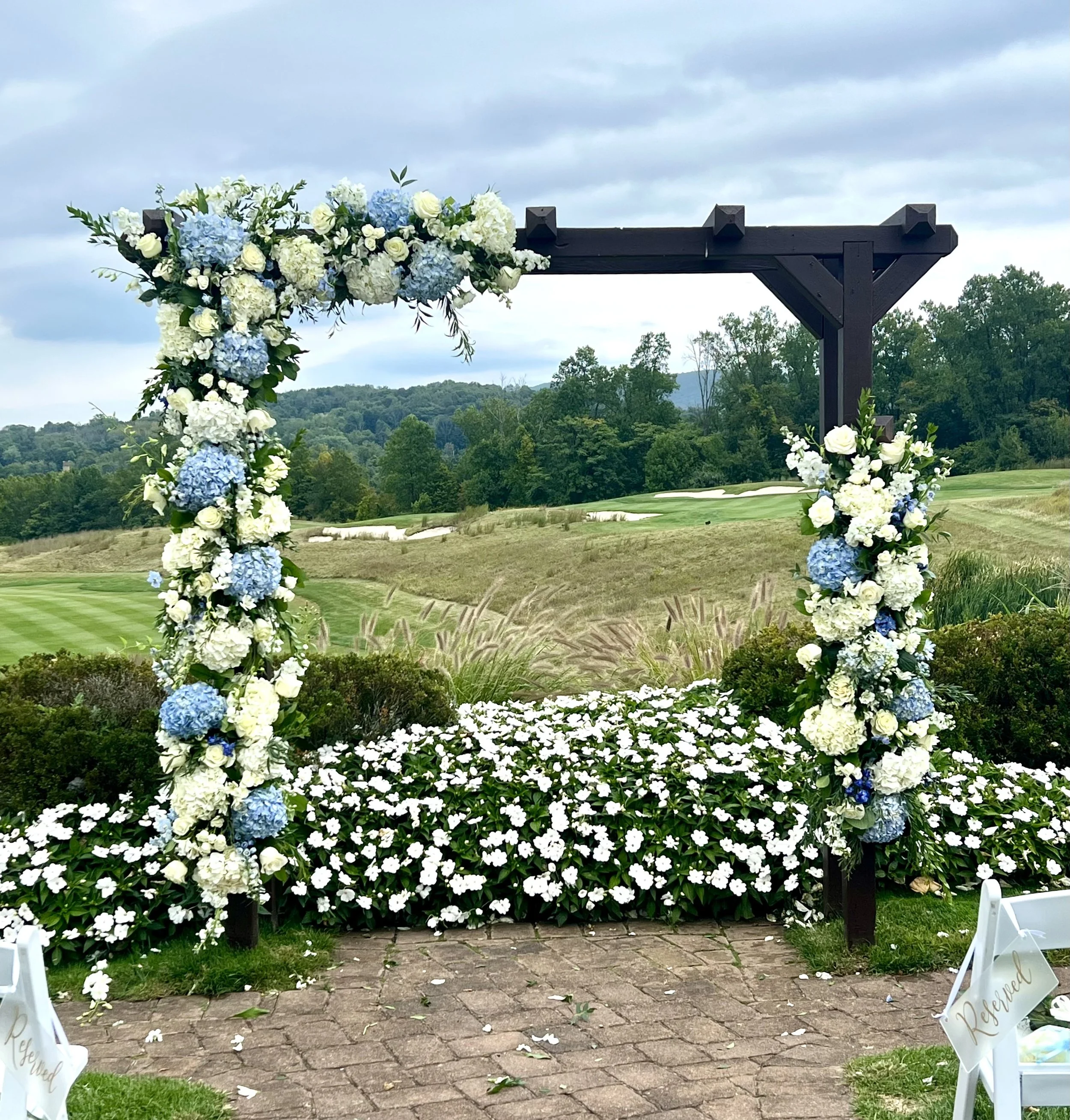 Wedding arch decorated with white and blue hydrangeas, roses, and greenery on a farm or outdoor field with hills and trees in the background.