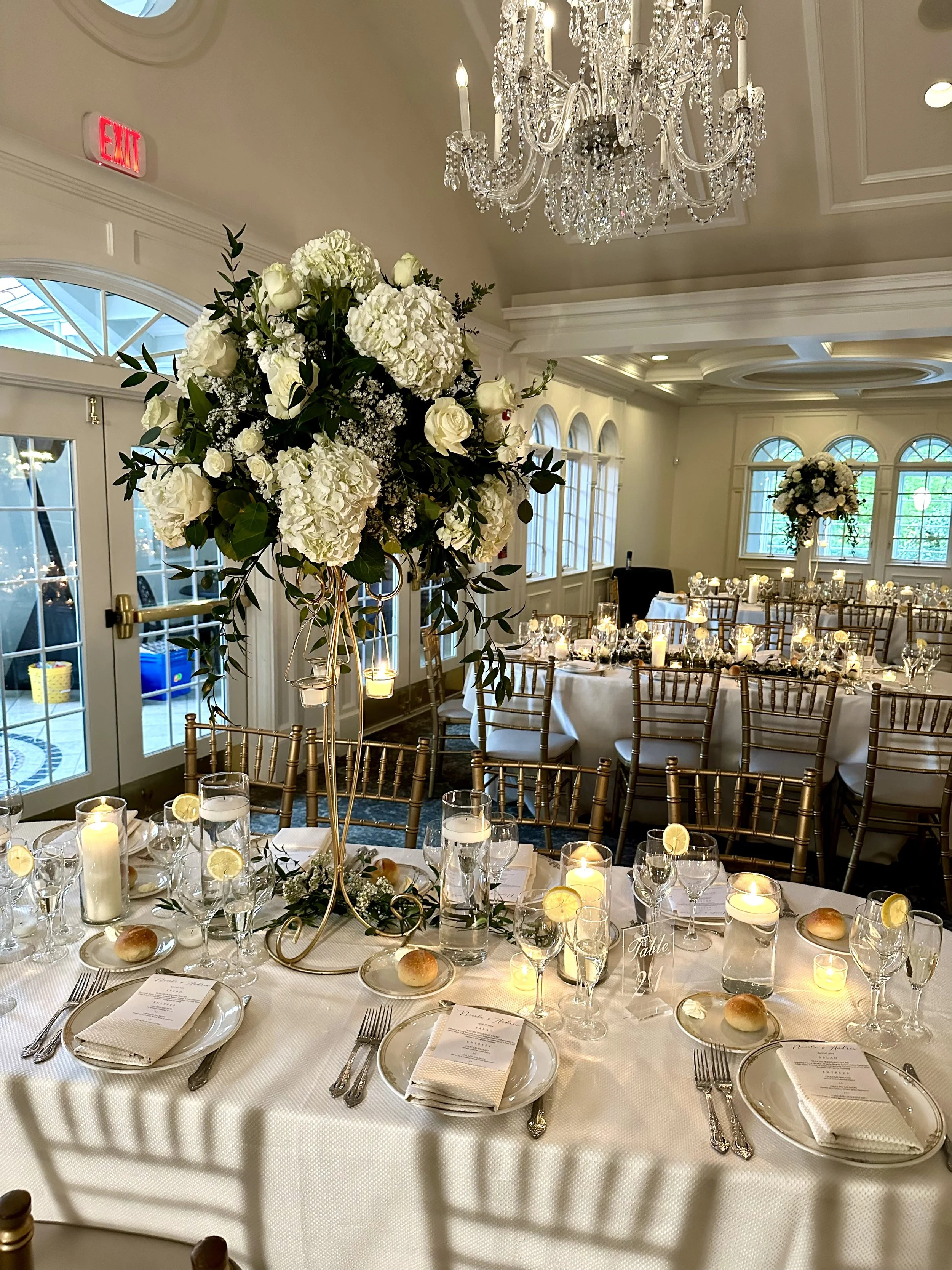 Elegant banquet table setup with white tablecloth, gold chairs, floral centerpieces, candles, and place settings with menus and dinner rolls in a luxurious room with a chandelier and large windows.