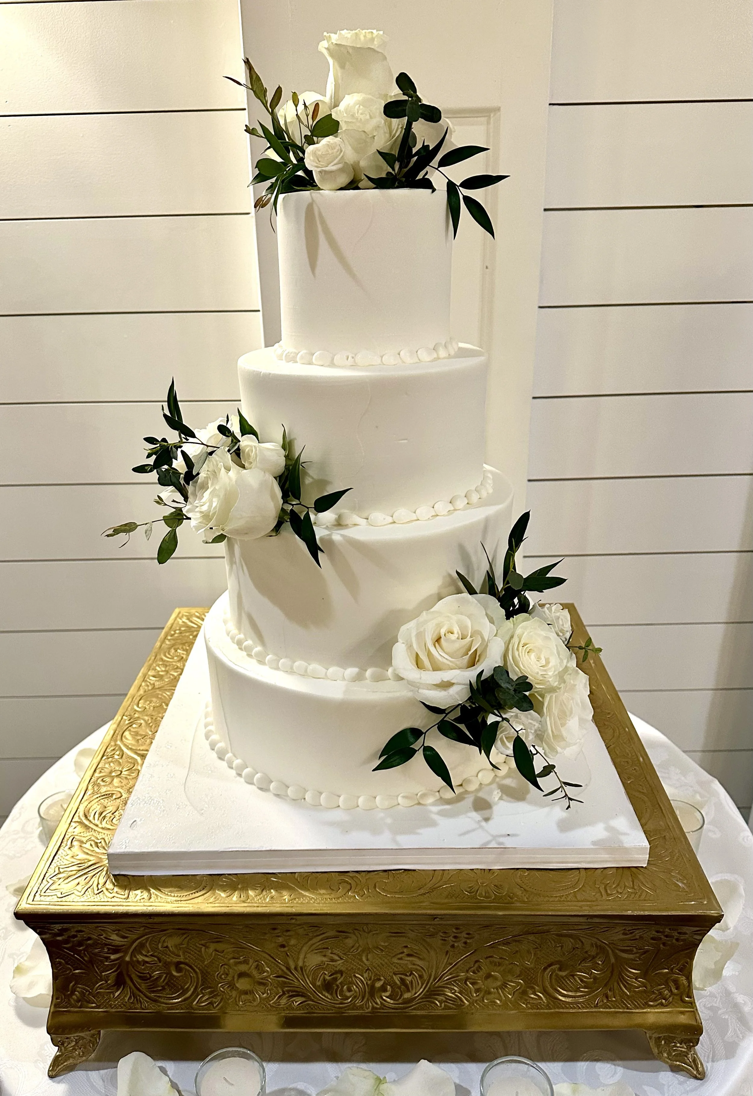 A four-tier white wedding cake decorated with white roses and green leaves, placed on a decorative gold-colored stand with intricate patterns.
