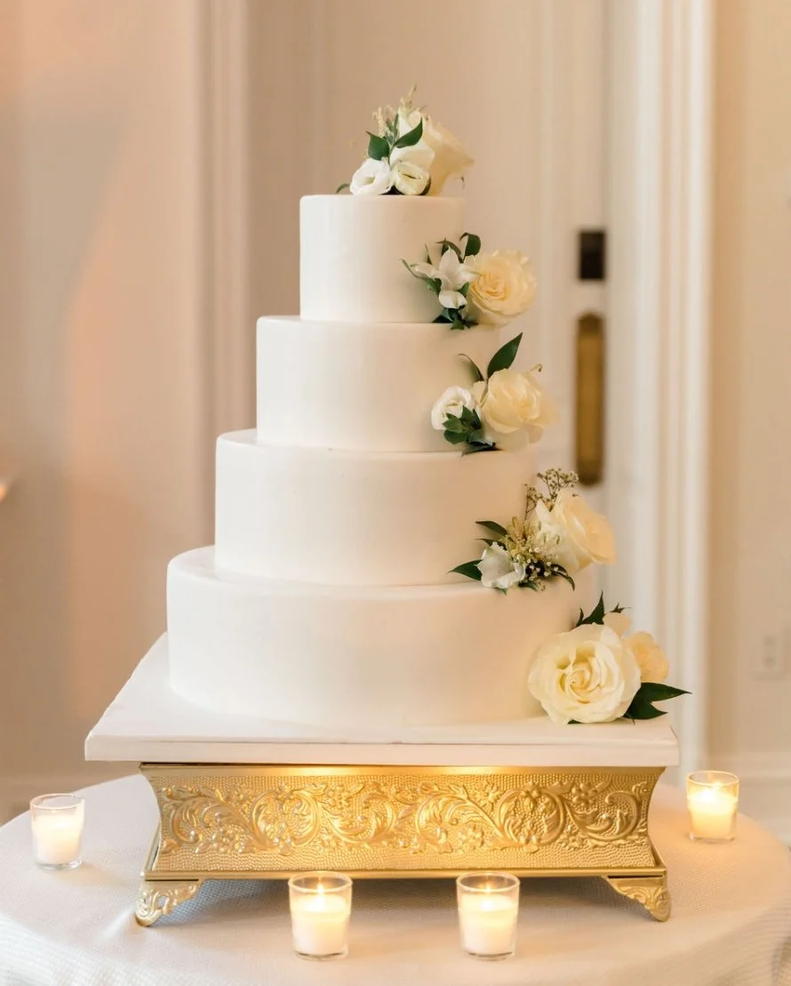 A four-tier white wedding cake decorated with white roses and green leaves, placed on an ornate gold stand surrounded by small lit candles.