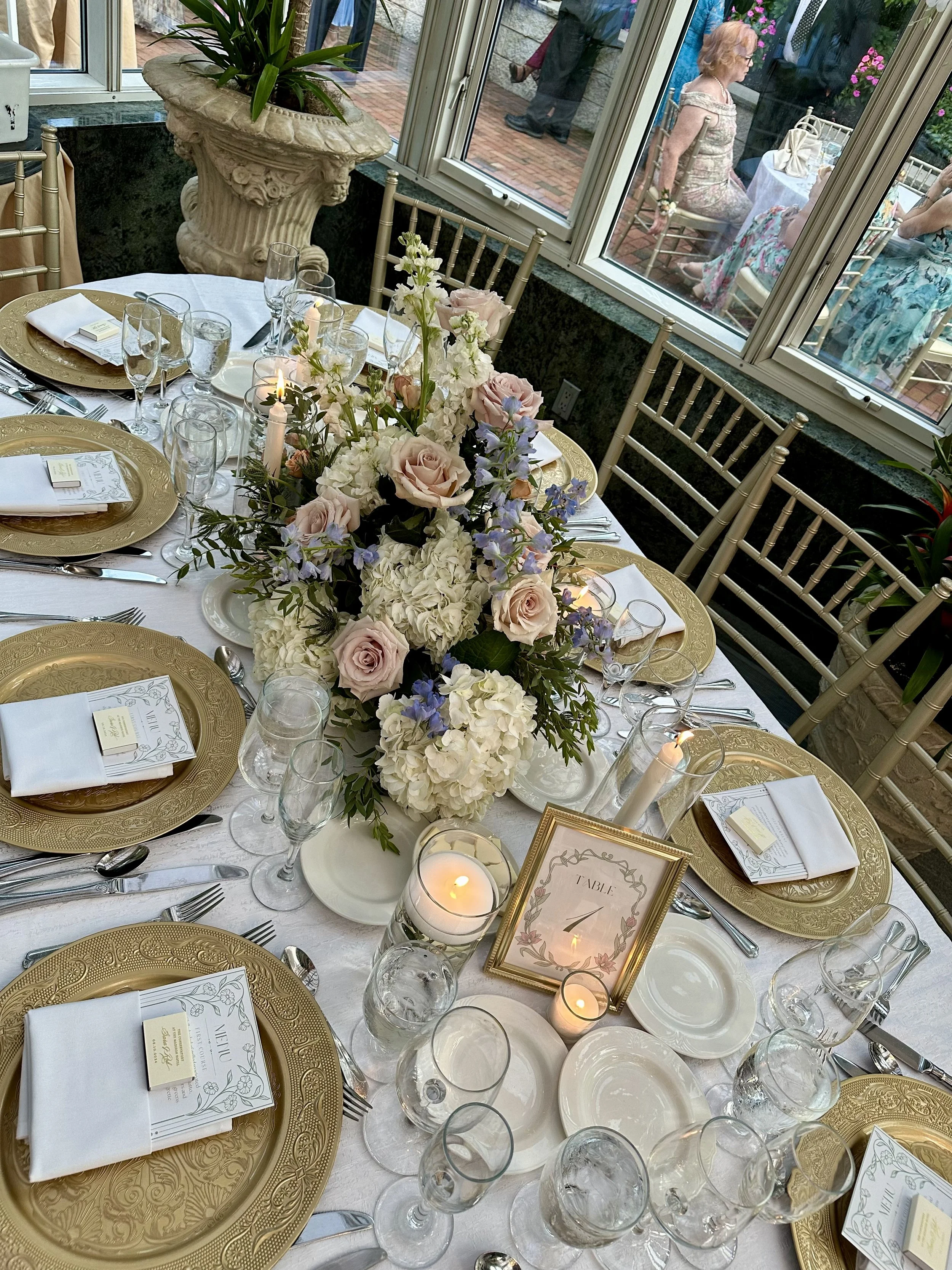 Elegant dining table set for a celebration with gold plates, white napkins, floral menus, and multiple glasses, topped with a centerpiece of pink and white flowers and candles. Outside through large windows, guests are mingling and sitting on chairs.