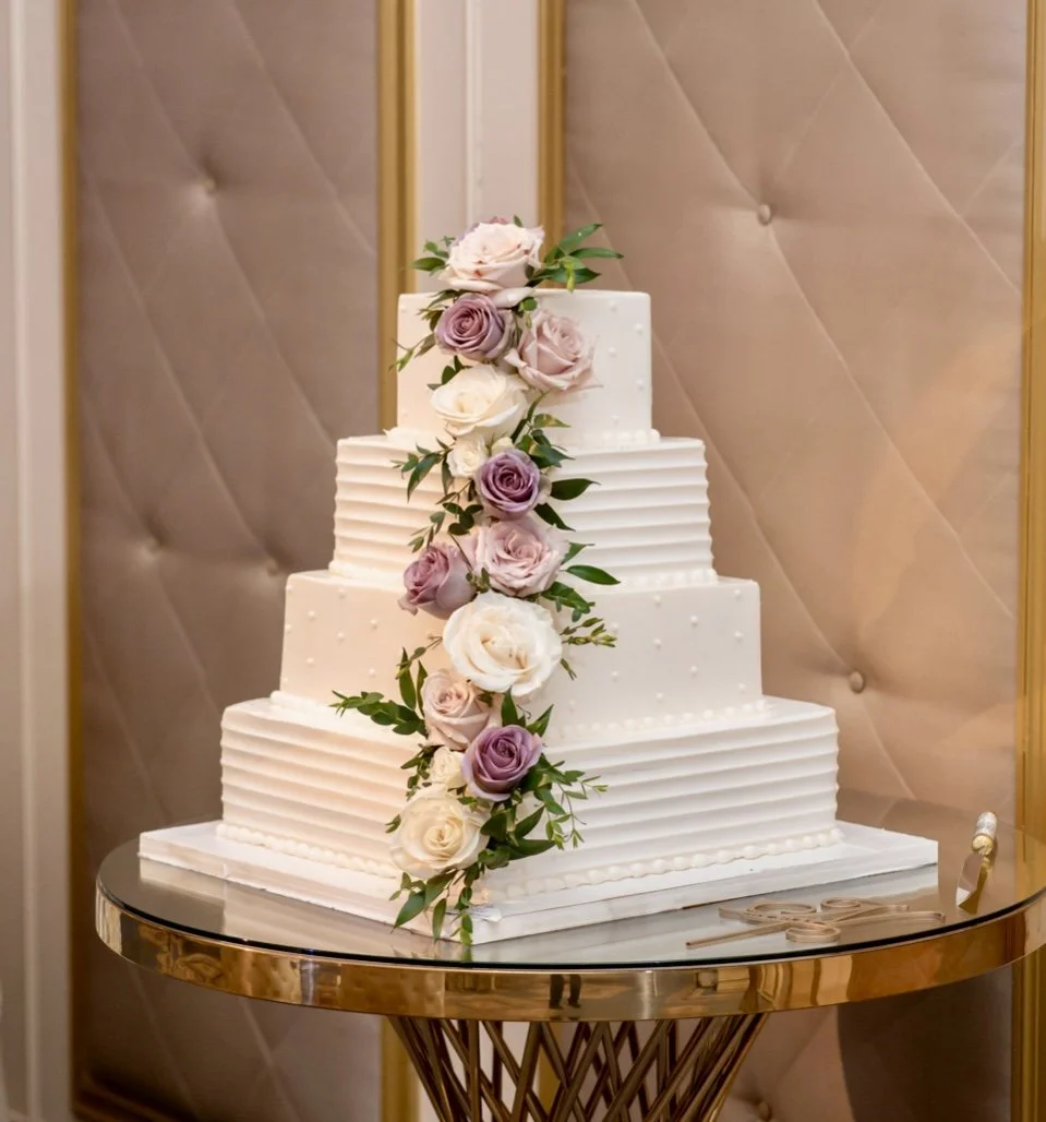 A four-tier white wedding cake decorated with cascading pink, purple, and white roses and green foliage, placed on a gold and glass table.