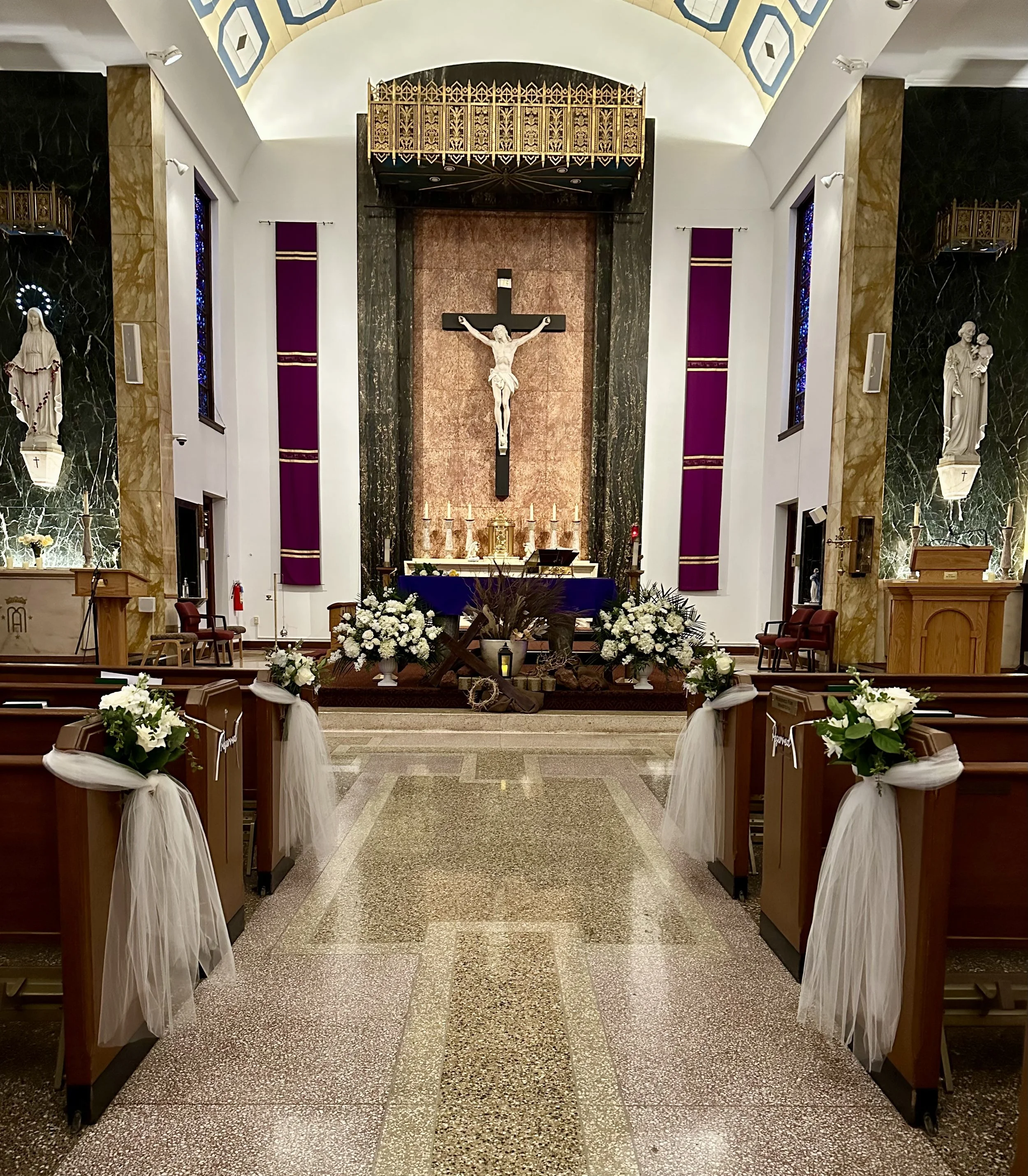 Inside a church, showing the altar with a crucifix of Jesus on the cross, decorated with flowers. The sanctuary has a polished stone floor, wooden pews decorated with white ribbons and flowers, and statues of saints on both sides. The background feat