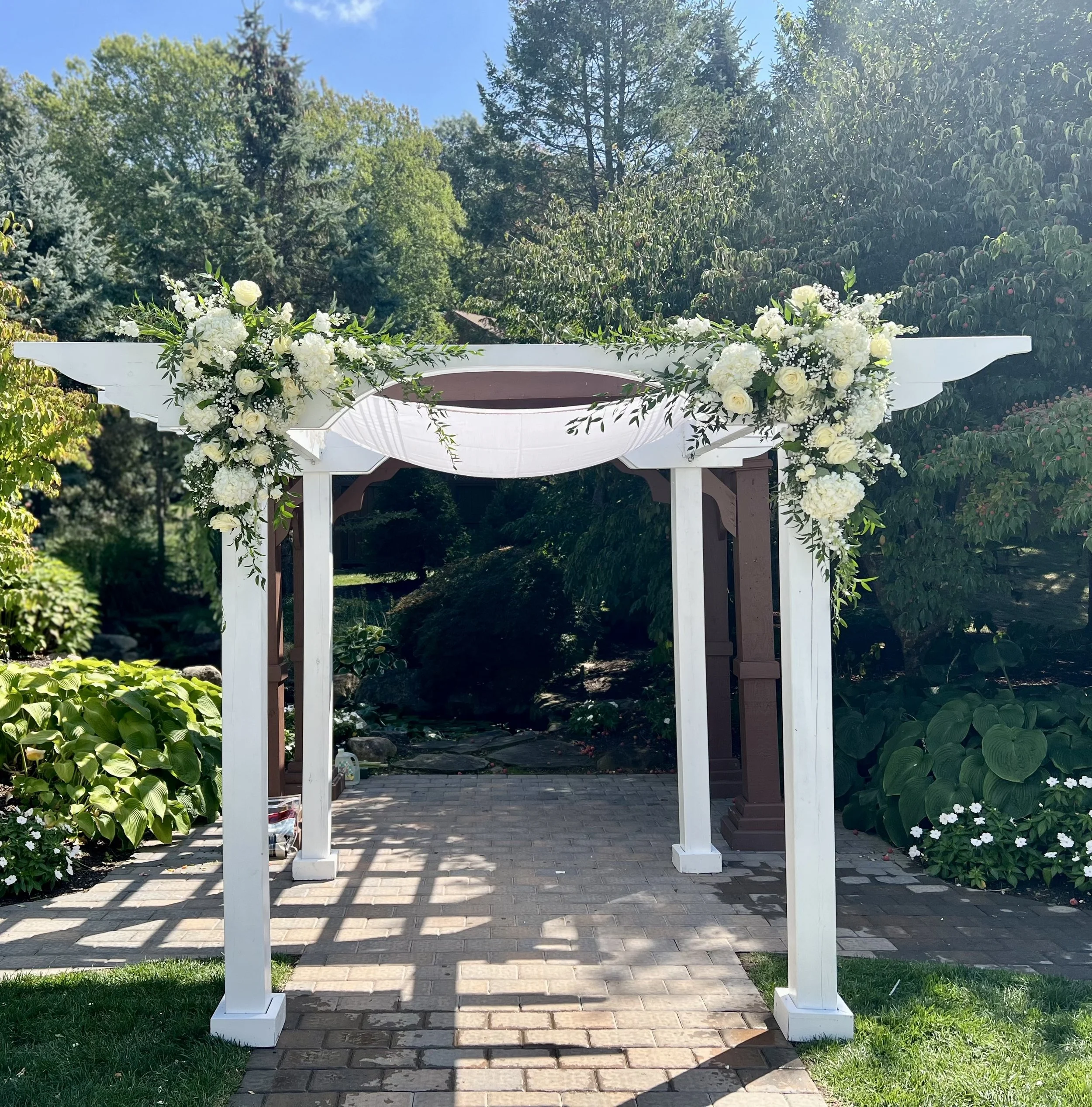 A white wedding arch decorated with white and light yellow flowers and greenery in an outdoor garden setting with a paved walkway, surrounded by lush green trees and plants under a blue sky.