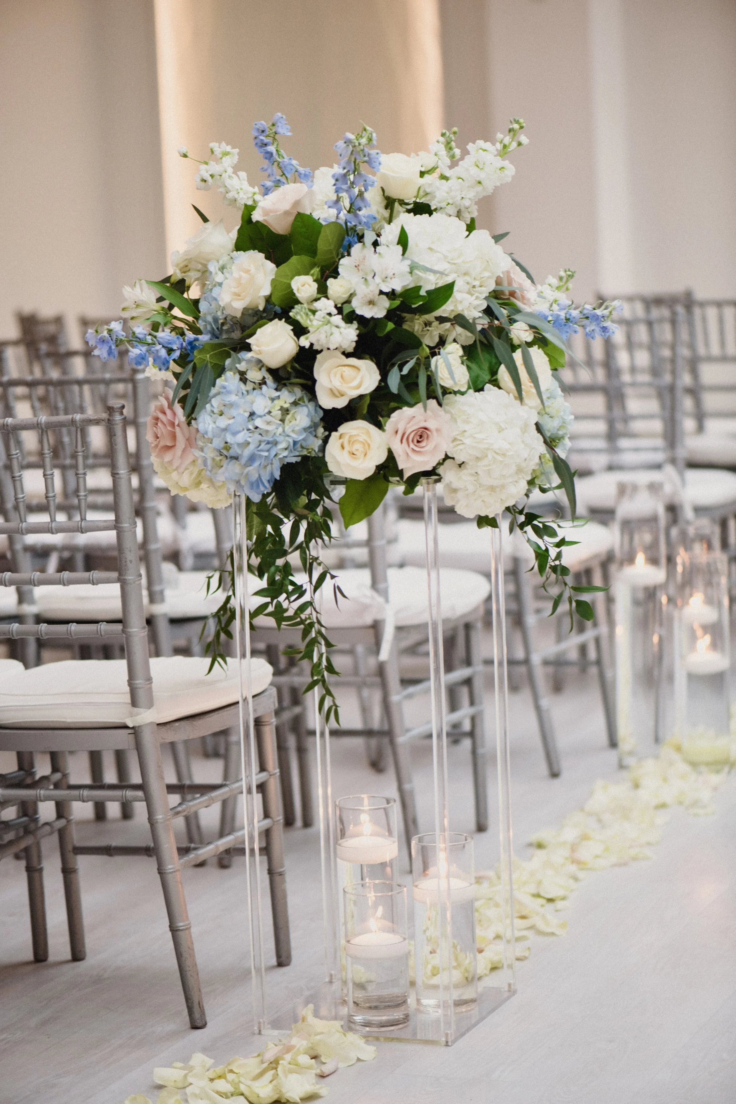 A floral centerpiece with white, pink, and light blue flowers on a tall, clear stand, surrounded by candles in glass holders on a white carpet with scattered flower petals at a wedding ceremony.