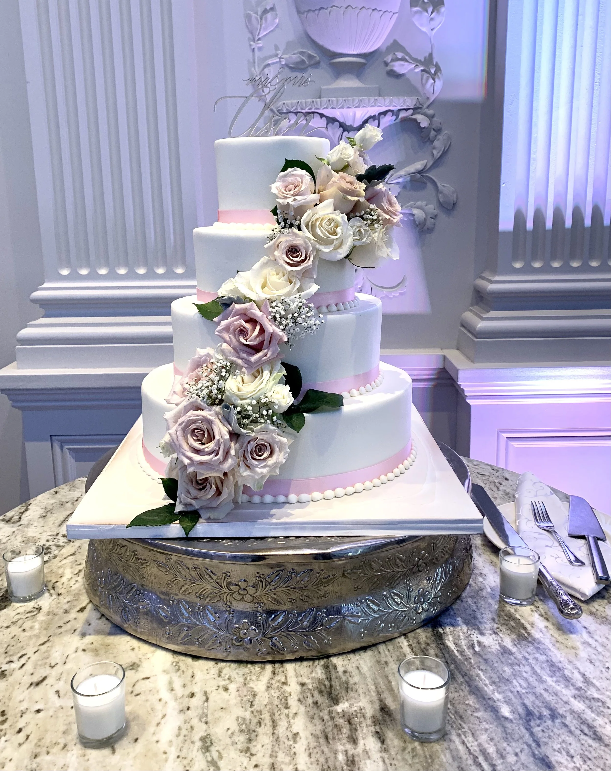 A multi-tiered wedding cake adorned with white and pale pink roses and baby's breath, placed on a decorative silver stand on a granite table, surrounded by small candles.