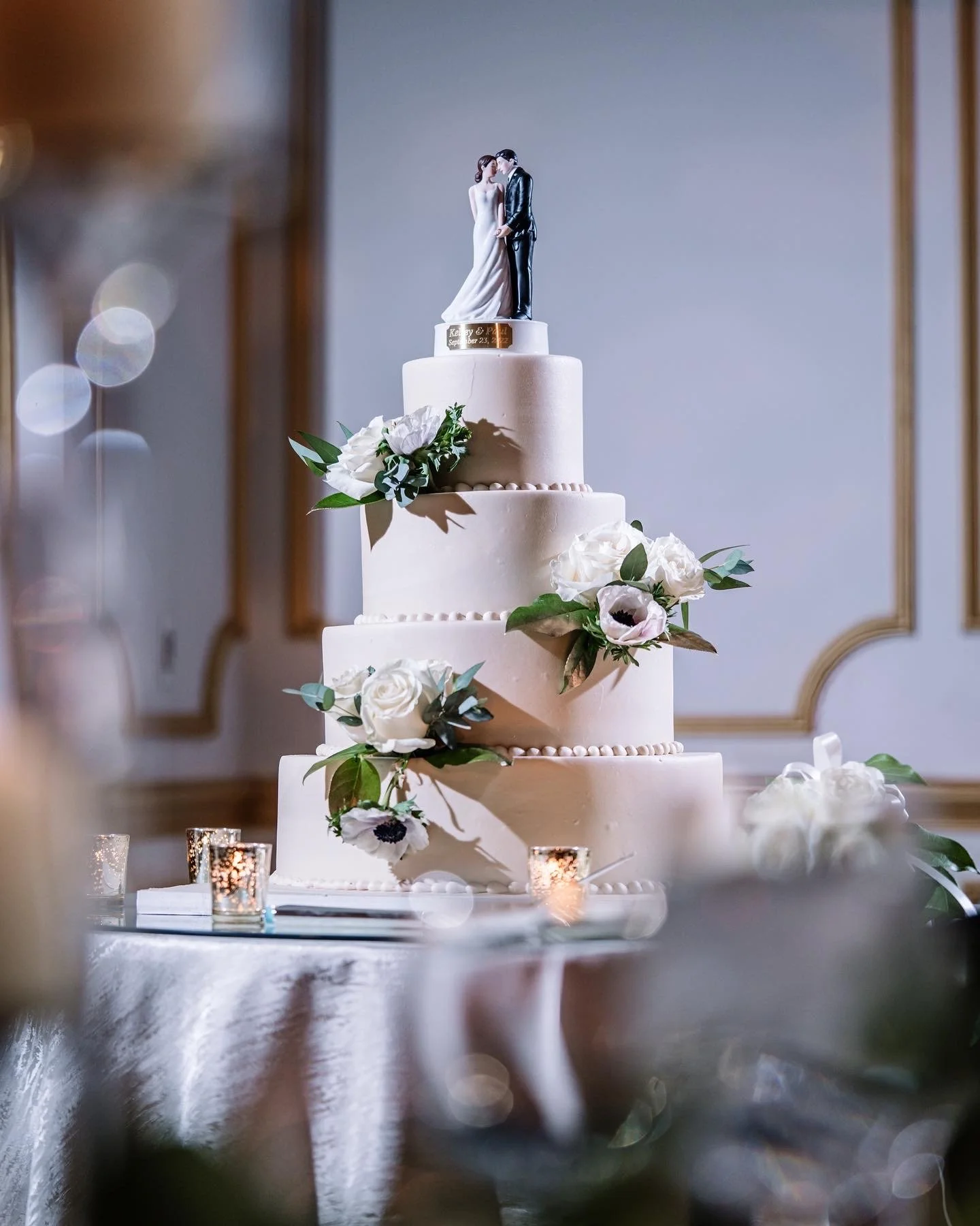 A multi-tiered wedding cake decorated with white and pink flowers and a cake topper depicting a bride and groom, on a table with candles and a reflective surface, in an elegant decorated room.