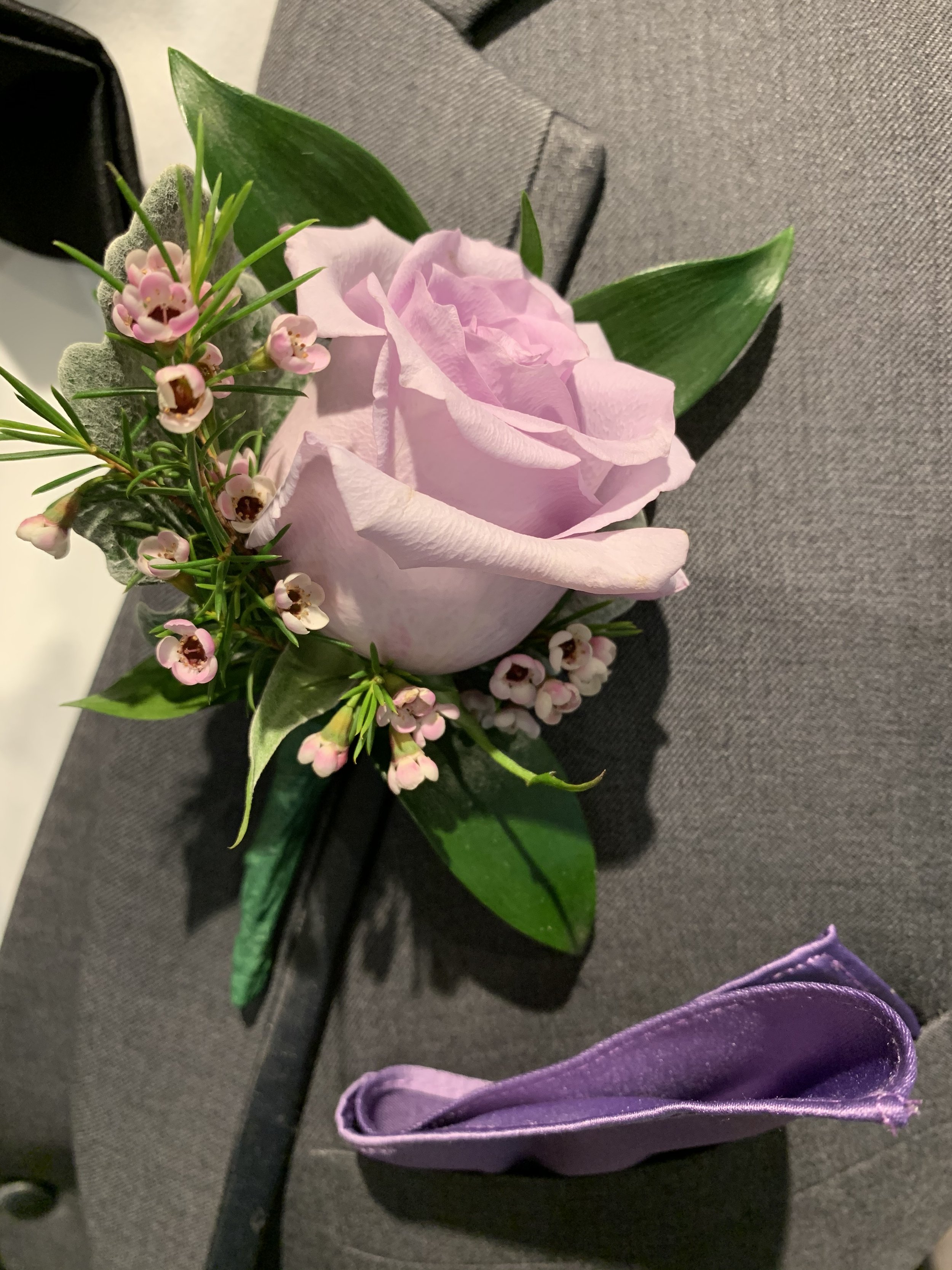 A boutonniere with a pink rose, pink and white small flowers, green leaves, and gray filler plant, attached to a gray fabric suit jacket, with a purple satin pocket square nearby.