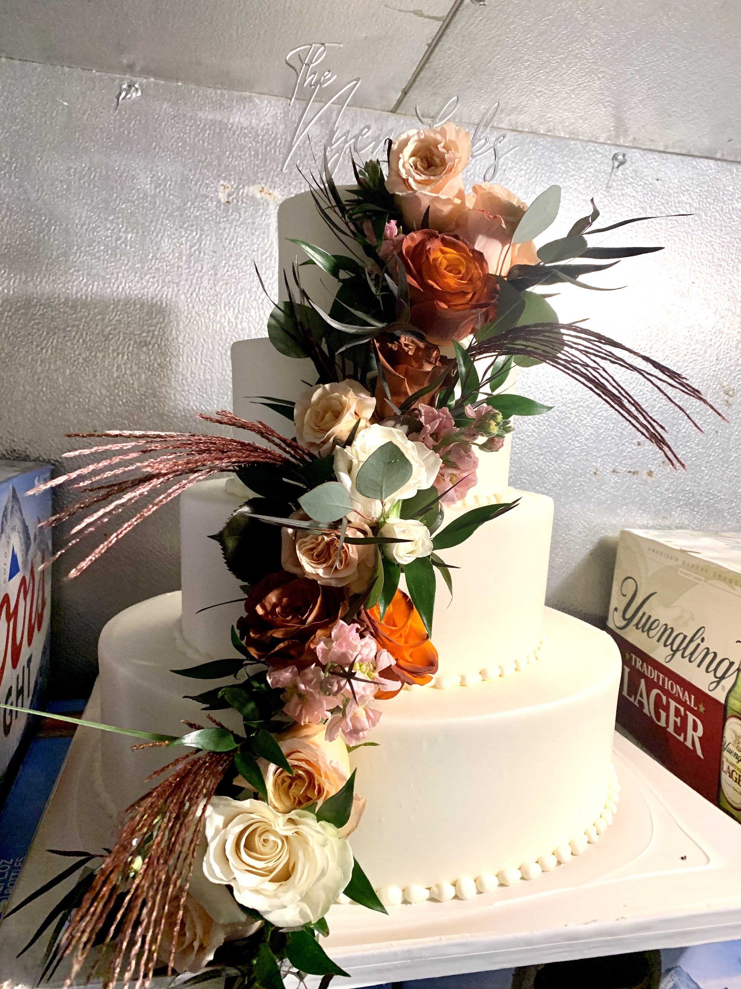 A white wedding cake decorated with a cascade of peach, cream, and orange roses, pink buds, eucalyptus leaves, and brown dried grasses, topped with a clear acrylic topper that says 'The Reyes'.