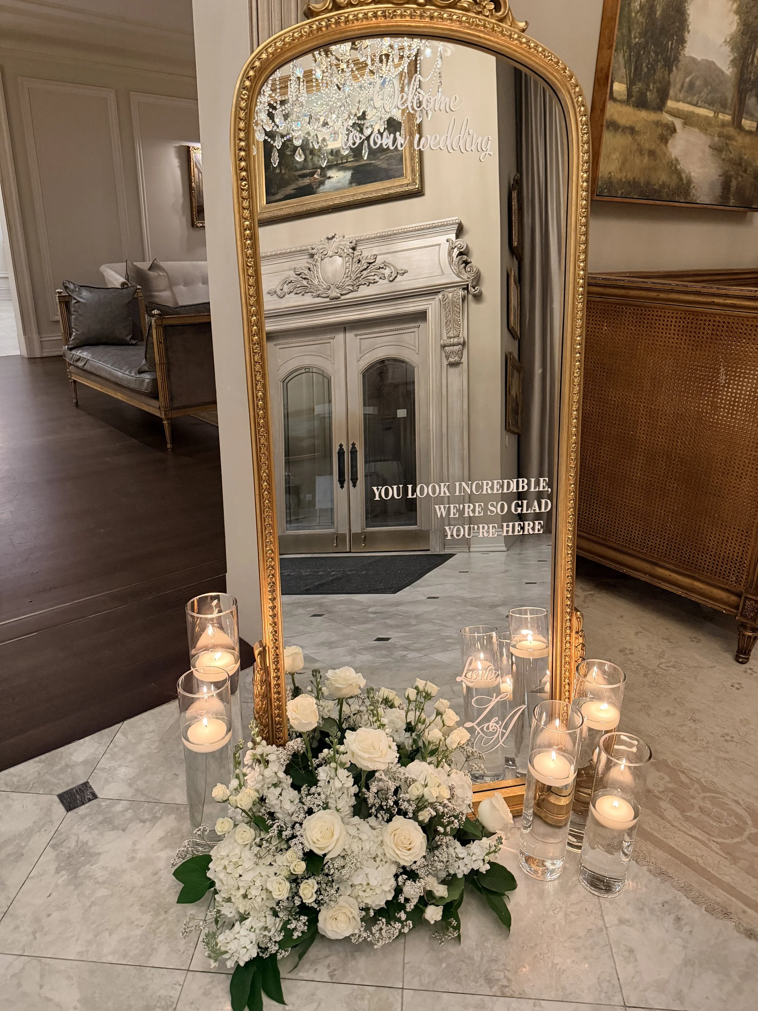 Decorative wedding display with a tall ornate gold-framed mirror, white floral arrangement with roses and baby's breath, illuminated candles in glass holders, and written message on the mirror that says, 'Welcome to our wedding. You look incredible, 