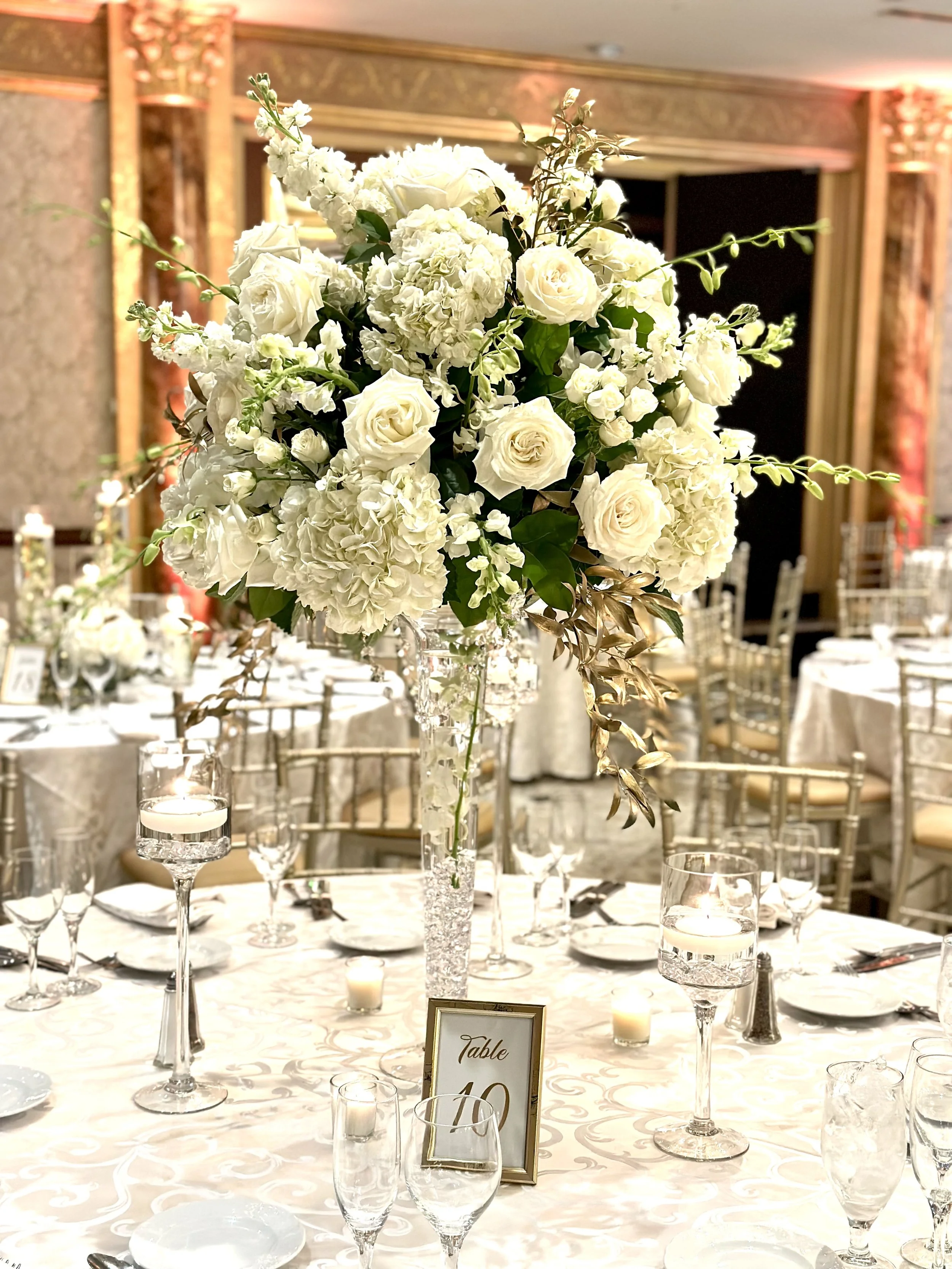 A large floral centerpiece with white roses, hydrangeas, and greenery on a table at a formal event venue with elegant decor and table settings.