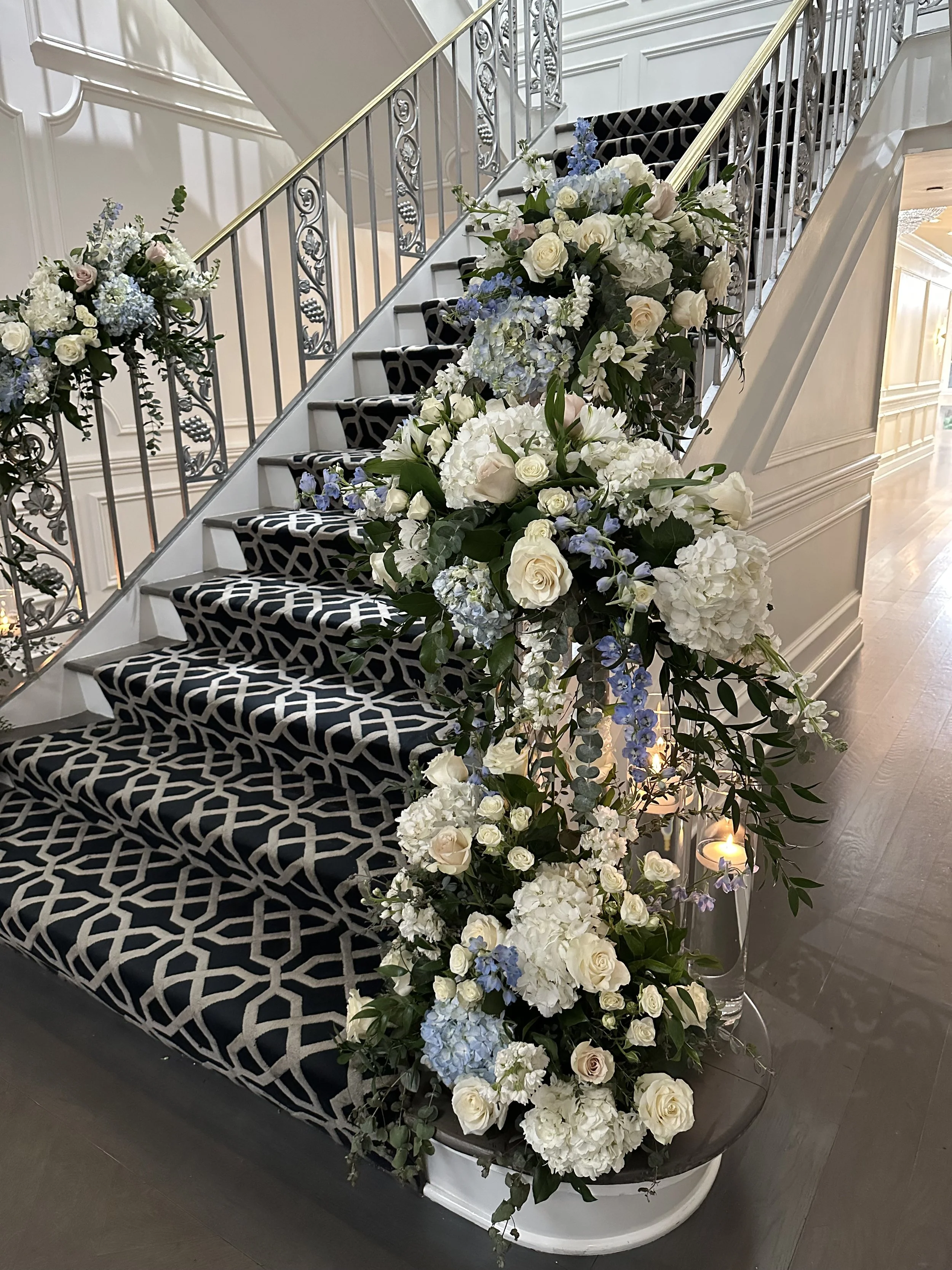 Elegant staircase decorated with cascading white and pale pink roses, white hydrangeas, blue delphiniums, and assorted greenery, with a lit candle in a glass holder at the base.