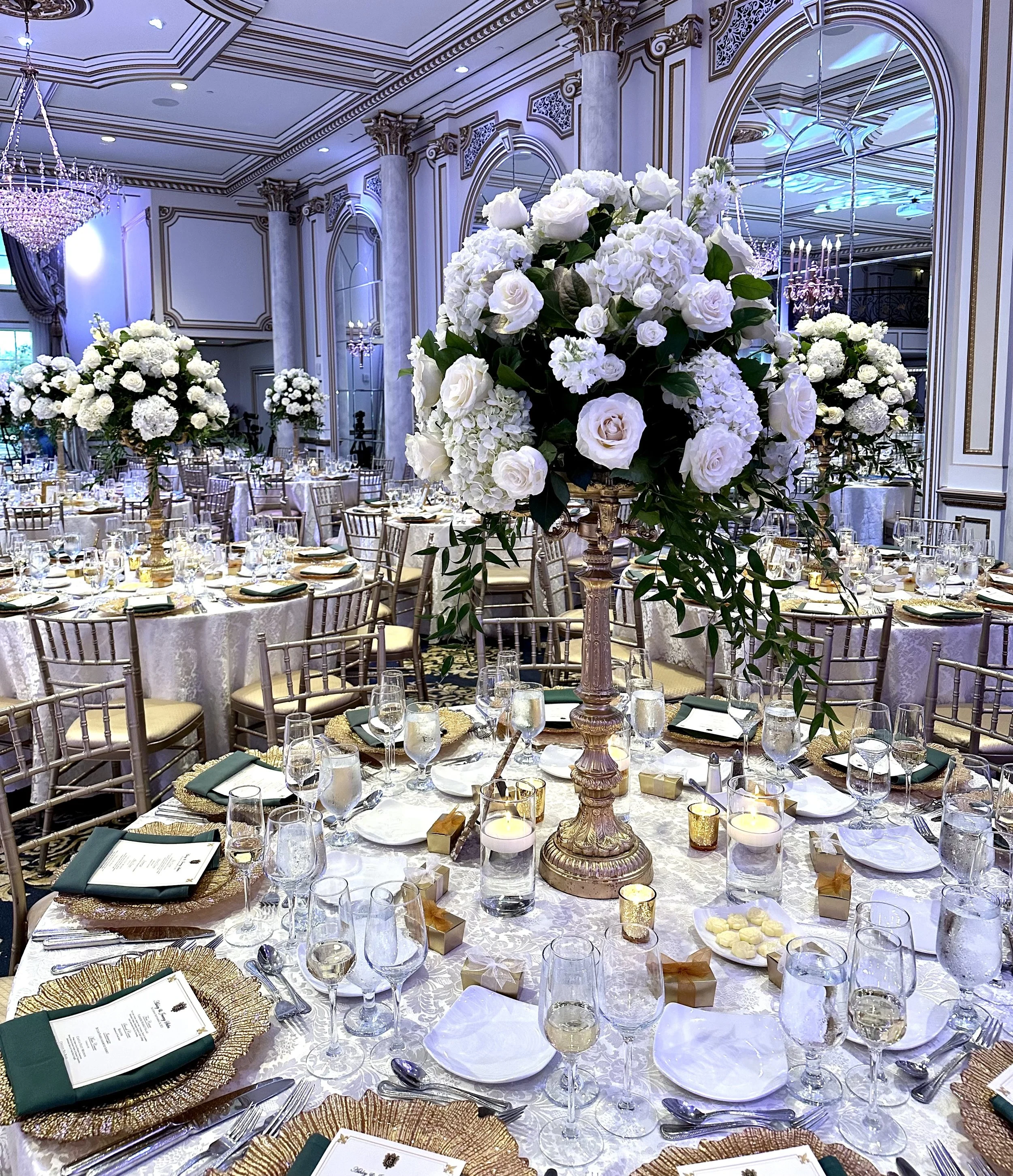 Elegant banquet hall decorated with multiple large floral arrangements of white roses and hydrangeas on golden stands, round tables with white tablecloths, place settings, and glassware, ornate molding and columns on the walls, large mirrors, crystal