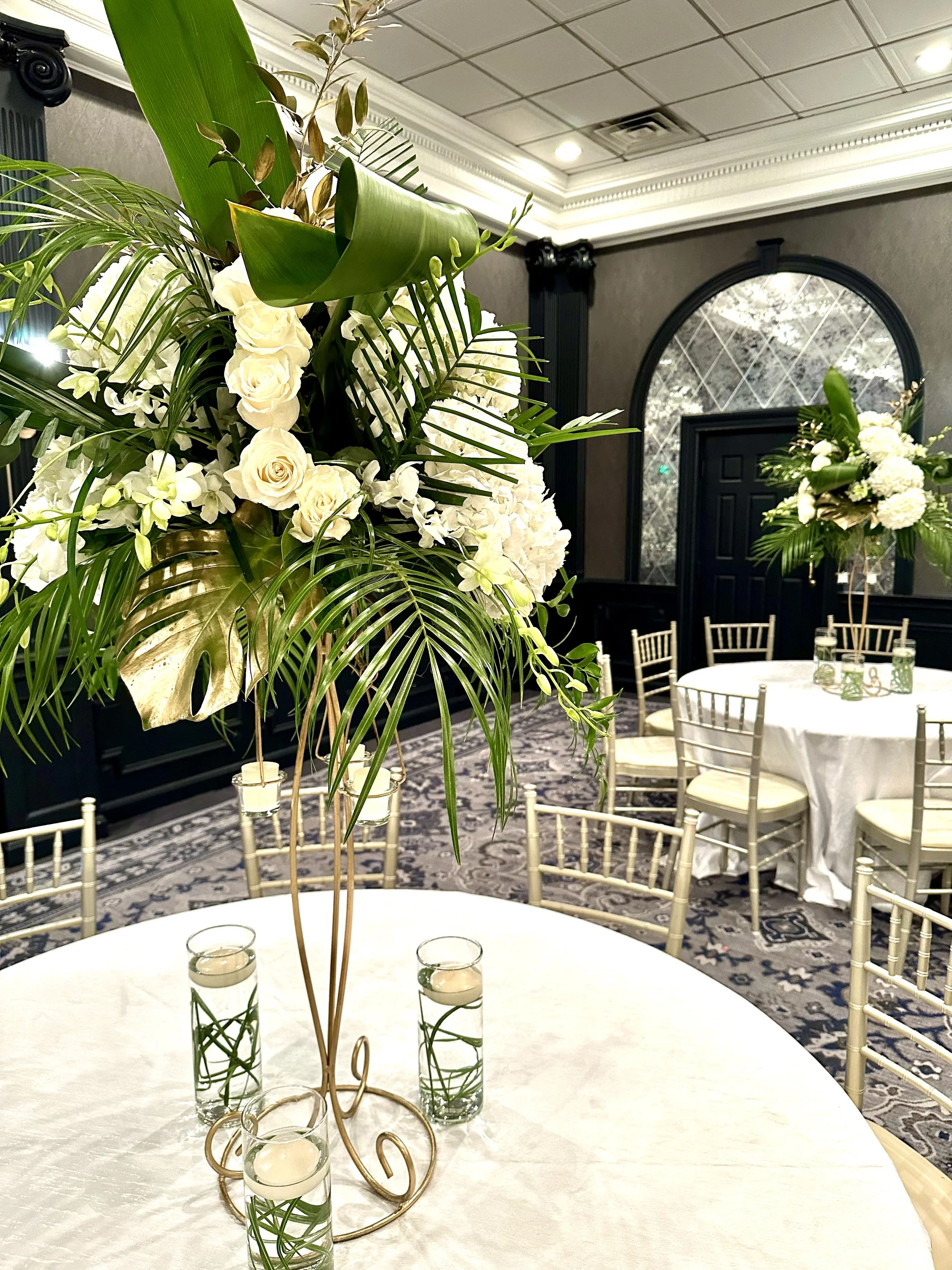 Elegant event room with white round tables, gold chairs, centerpieces of white flowers and tall green foliage, and candles in glass holders.