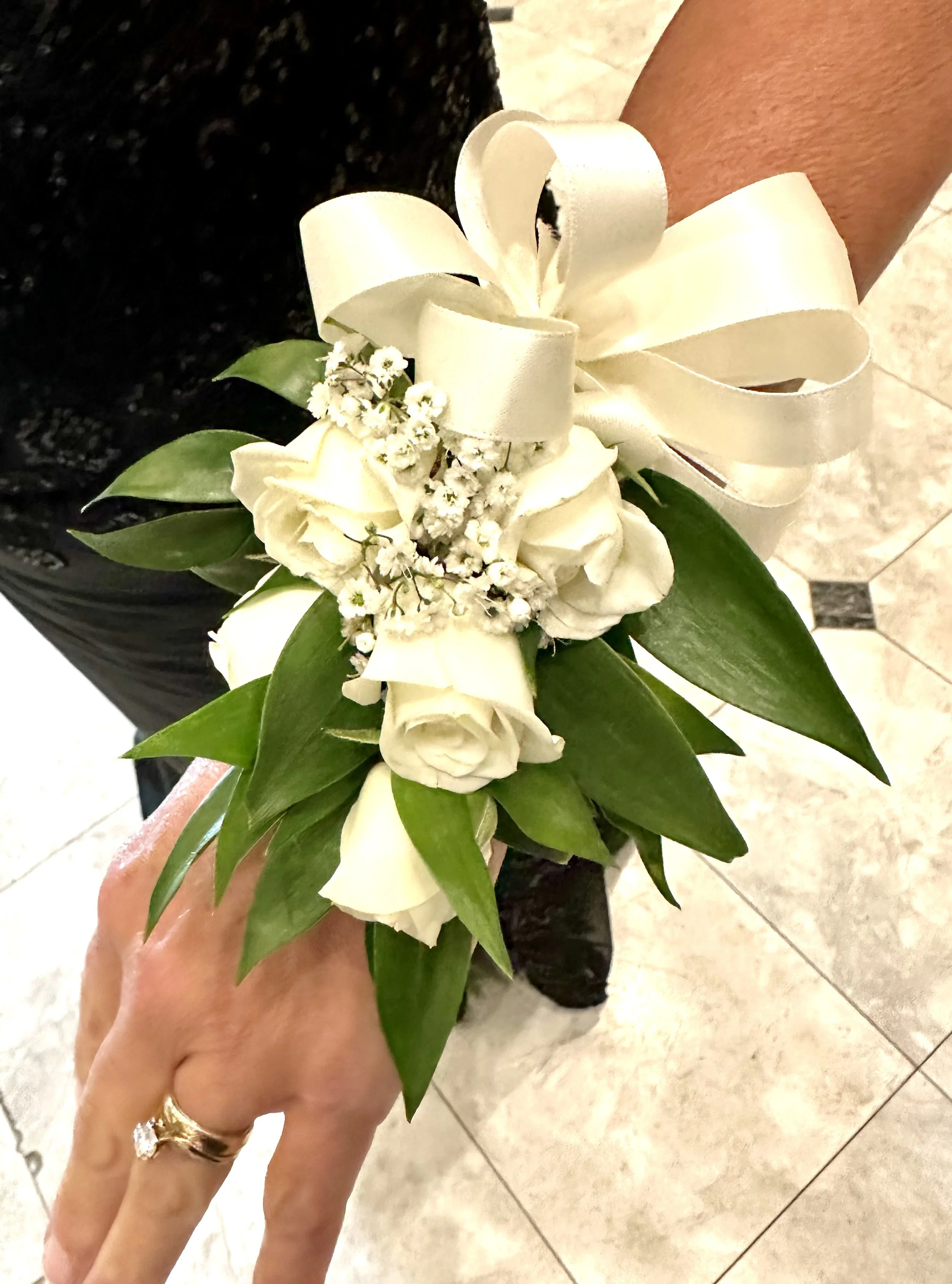 Close-up of a floral corsage held by a person's hand, featuring white roses, small white filler flowers, green leaves, and a cream-colored satin ribbon bow.
